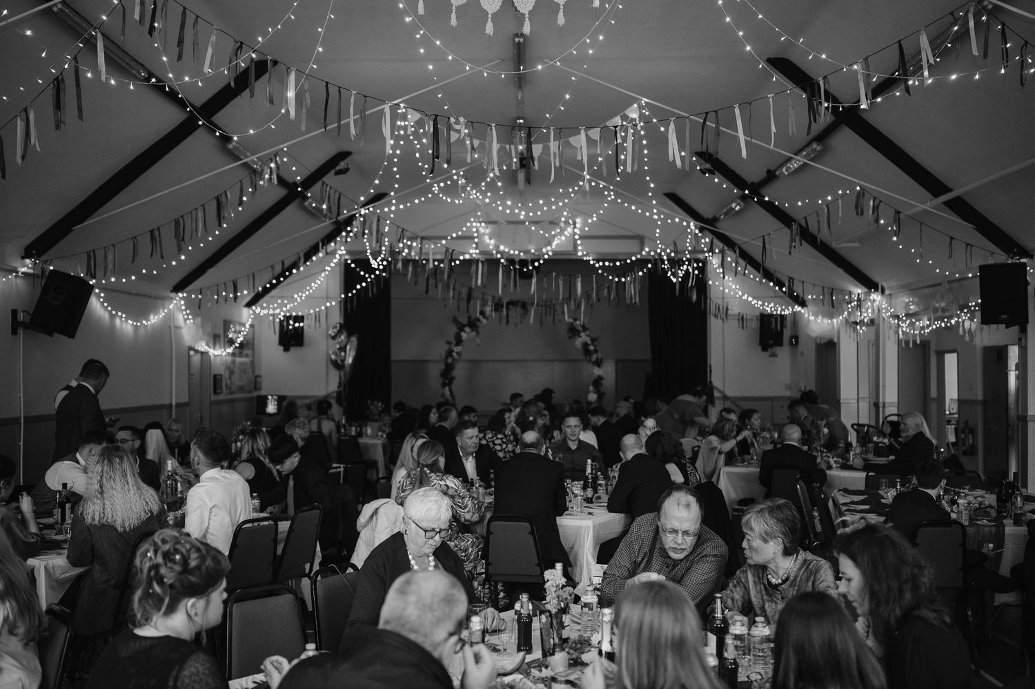 Large wedding reception hall decorated with hanging lights and guests seated at tables.