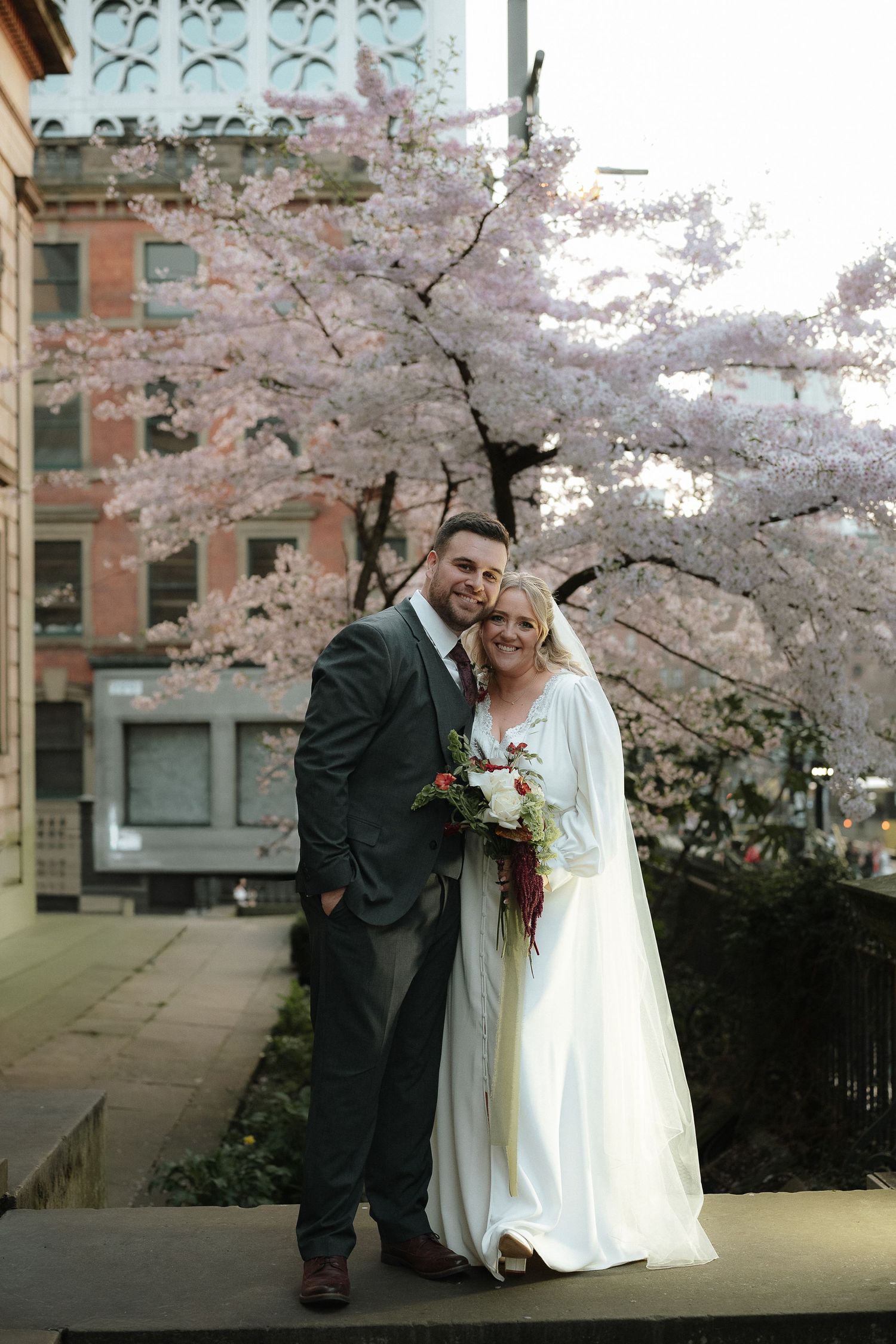Bride and groom pose before blooming cherry blossom trees in an urban setting, bride holding a bouquet. Manchester Wedding Photographer