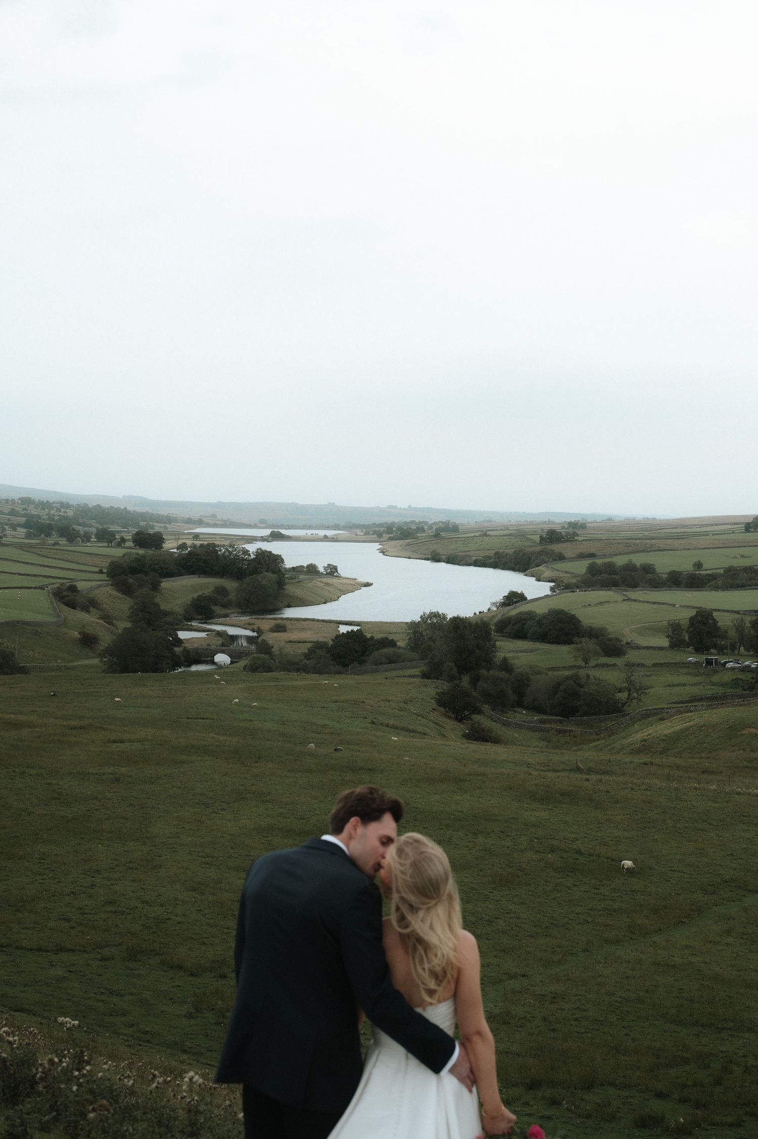 Newlyweds embrace while overlooking a scenic river valley landscape on their wedding day. Blackton Grange Wedding. Luke Hassall Photography.