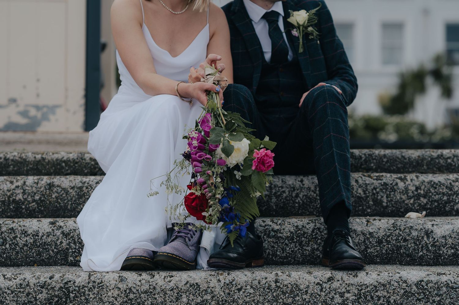 Wedding couple sitting on stone steps with colorful wedding bouquet and elegant attire.