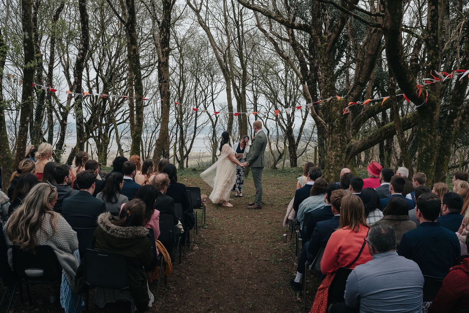 Outdoor forest wedding ceremony with guests seated along wooded path.