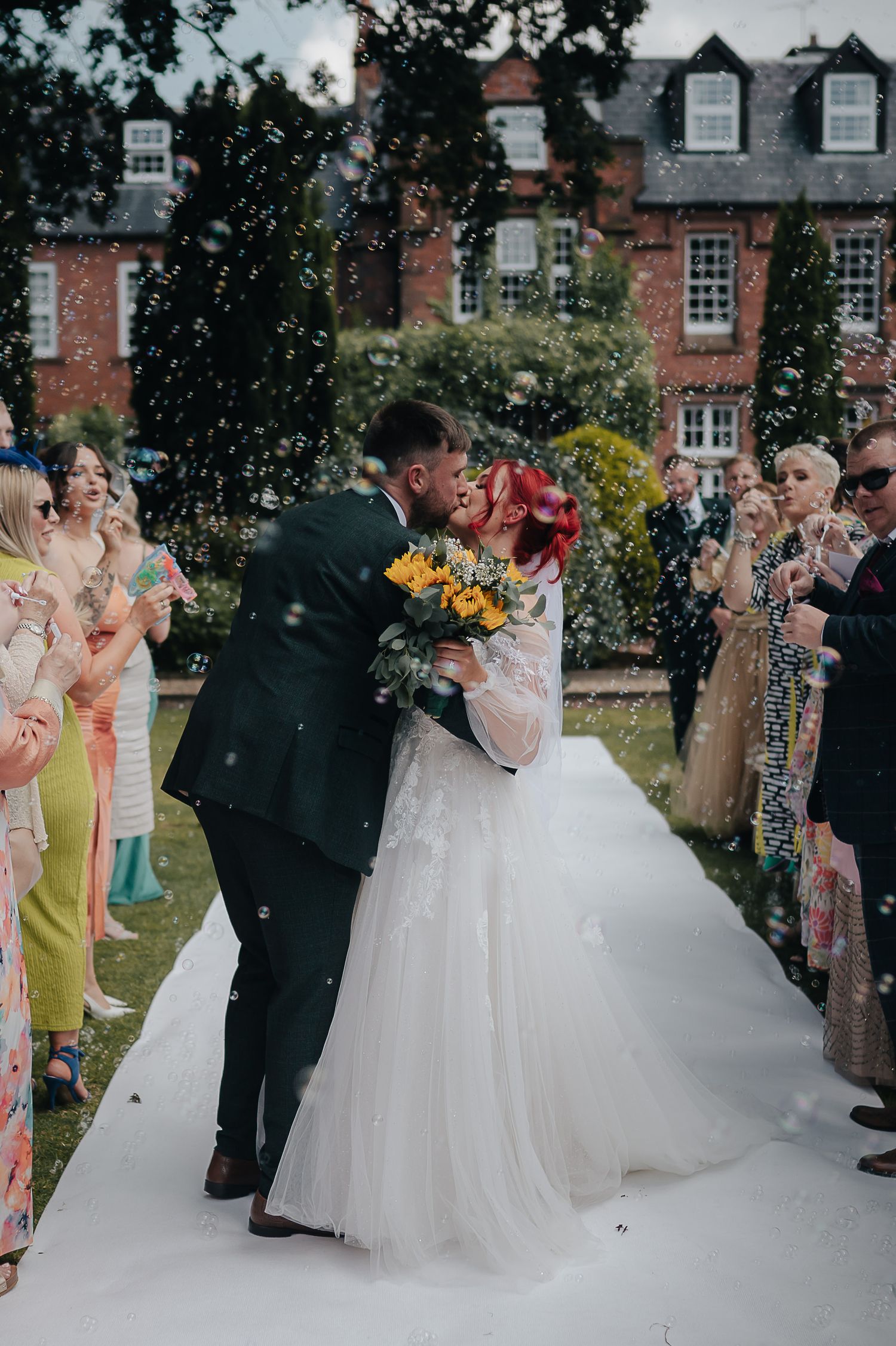 Wedding couple sharing their first kiss on a white aisle runner while guests celebrate with confetti.