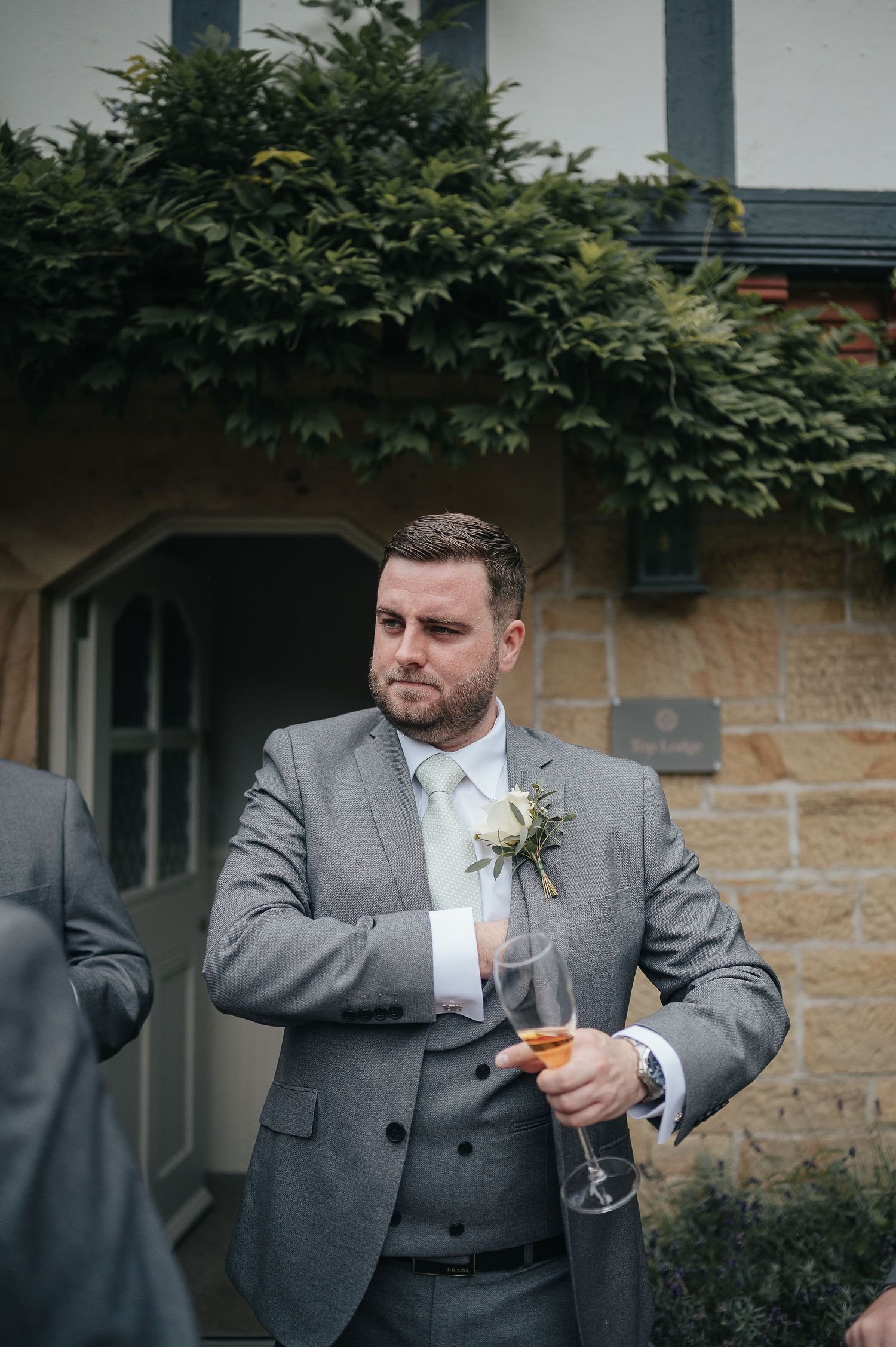 Wedding guest in grey suit holding champagne glass outside Tudor-style building.