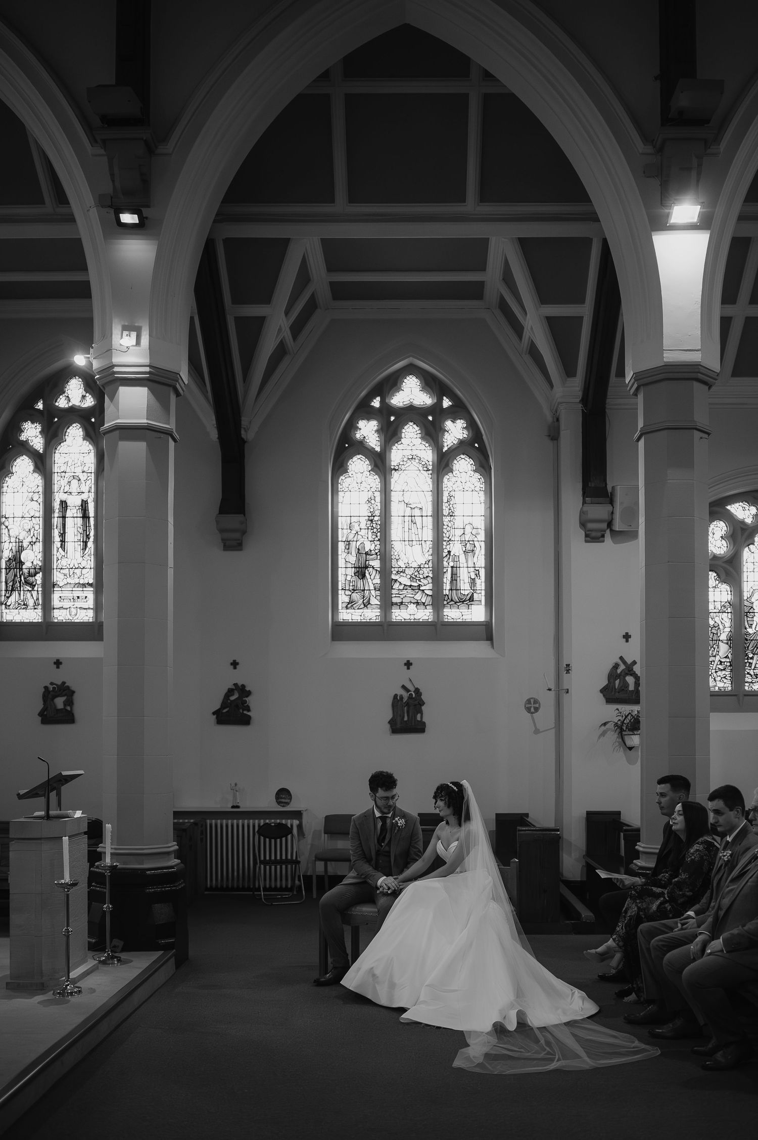 Black and white photo of a wedding couple seated in a church with stained glass windows and Gothic architecture.