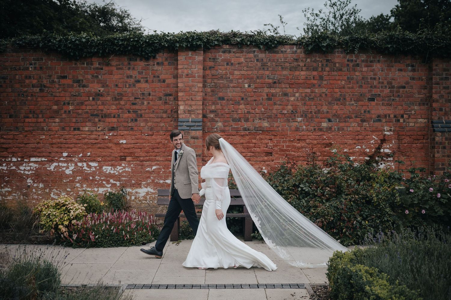 Wedding couple walking along brick garden wall with flowing white veil trailing behind.