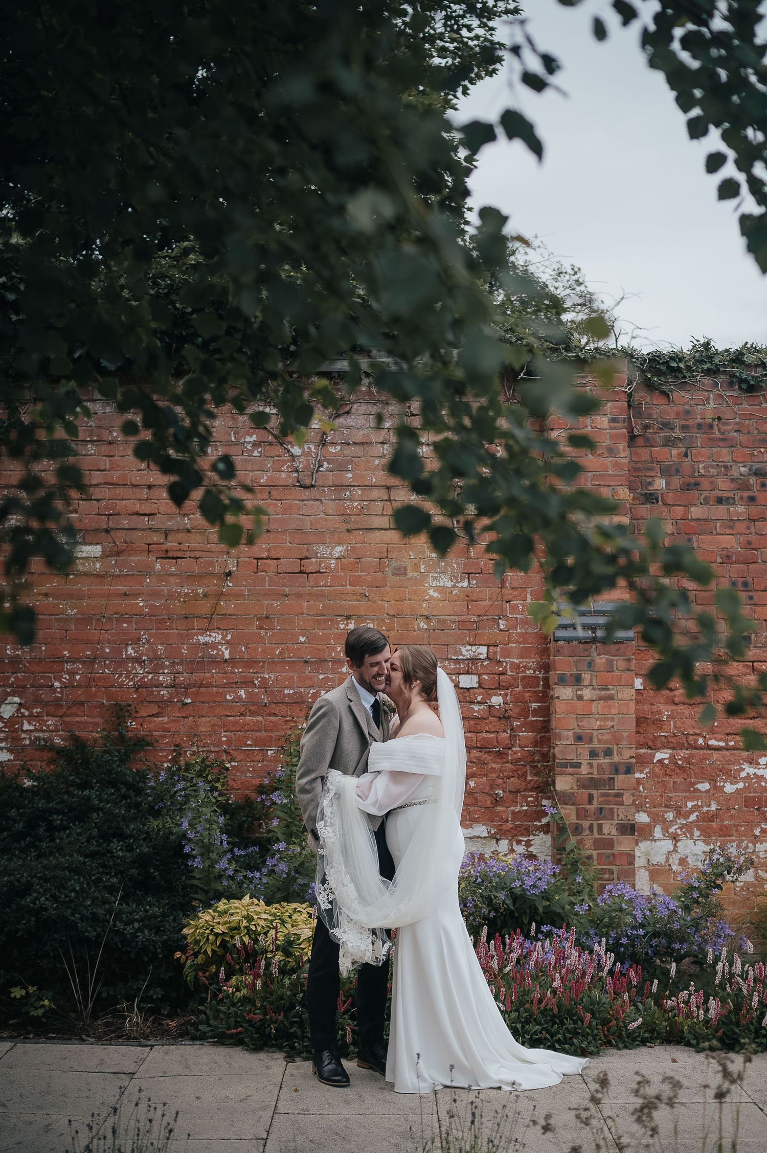 Romantic wedding portrait against rustic brick wall with blooming garden flowers.