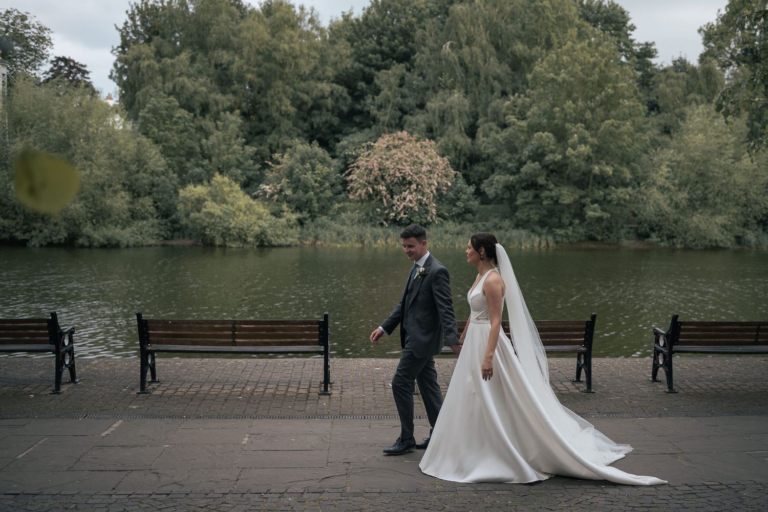A couple strolls along a lakeside path with benches and autumn trees in the background.