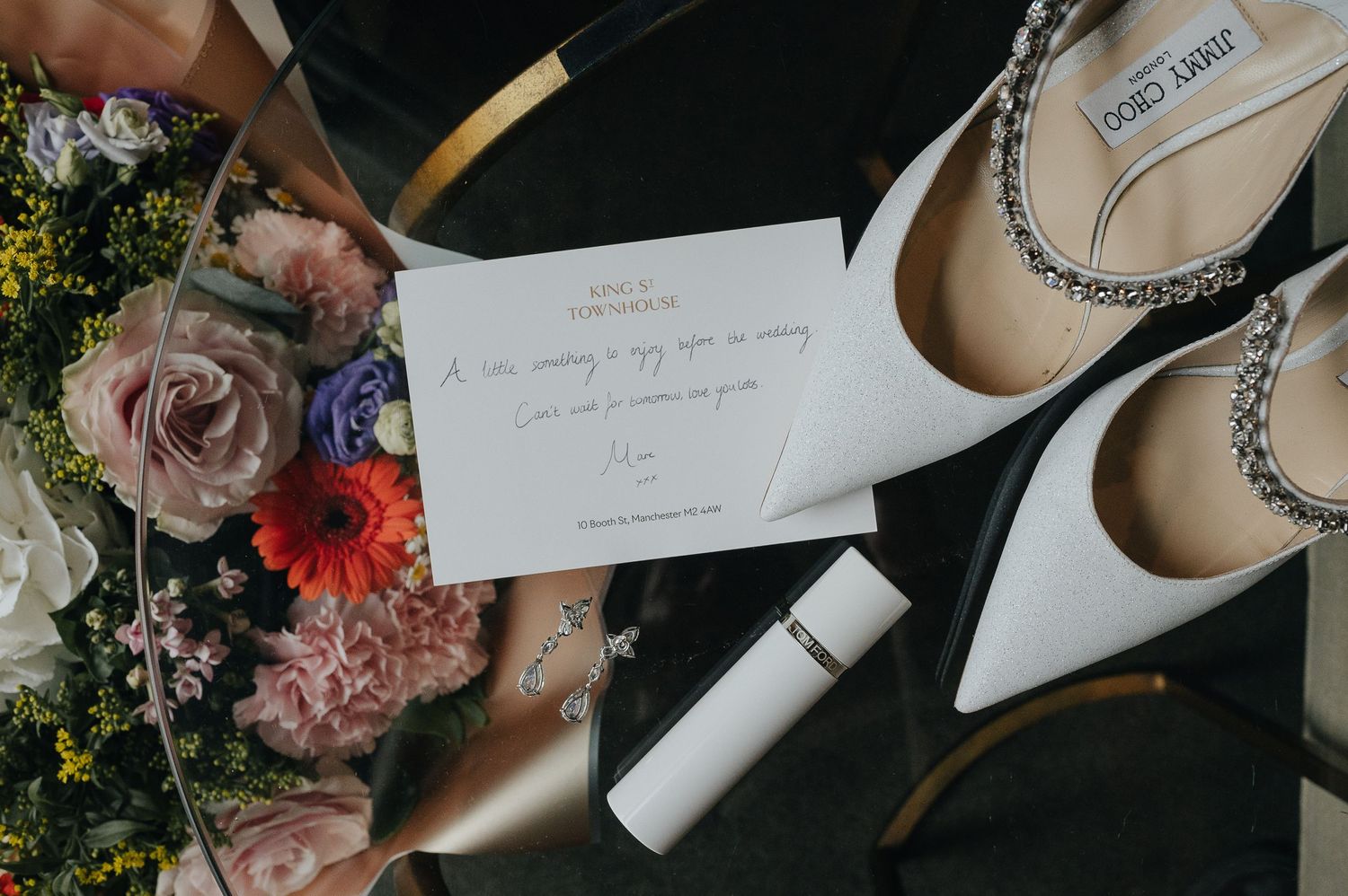 Wedding details flatlay with white shoes, handwritten note, and pink floral arrangements.