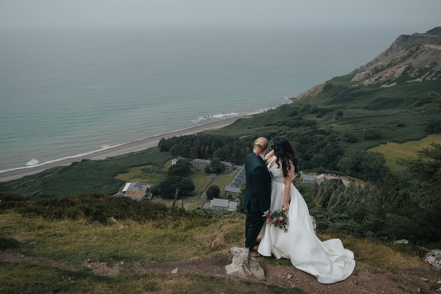 Wedding couple stands on coastal cliffside with dramatic ocean views in background.