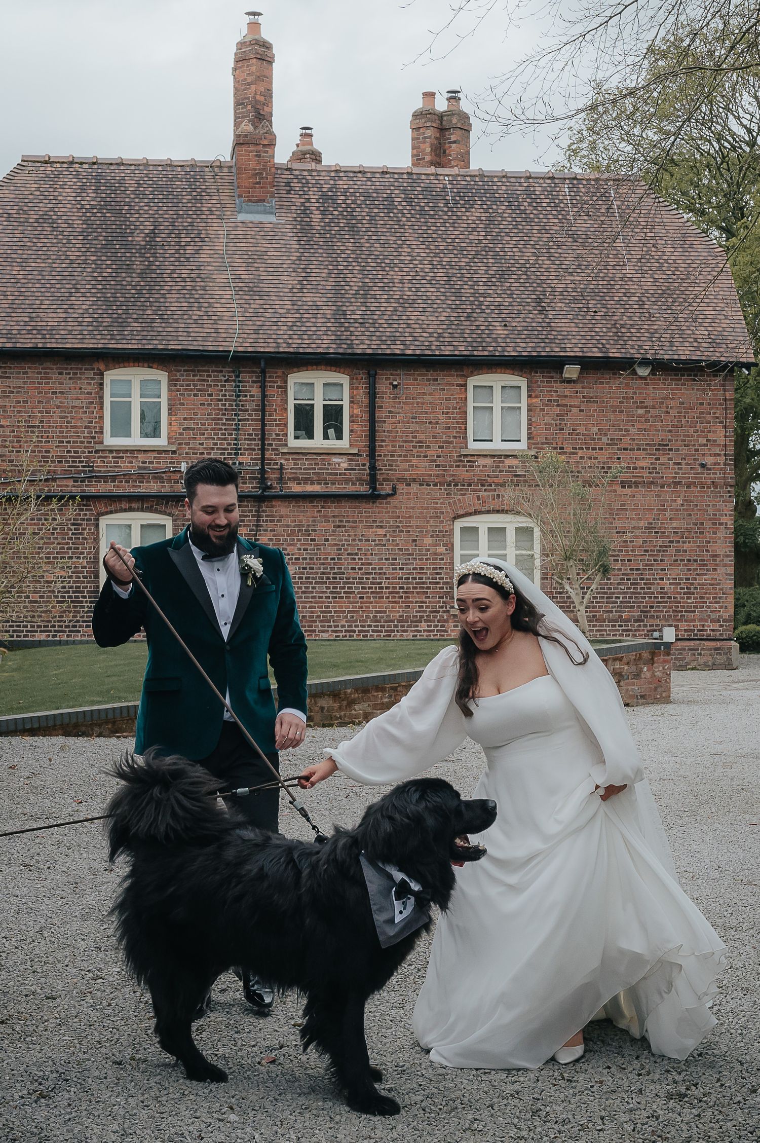 Newlyweds posing with their large black dog outside a brick country house.