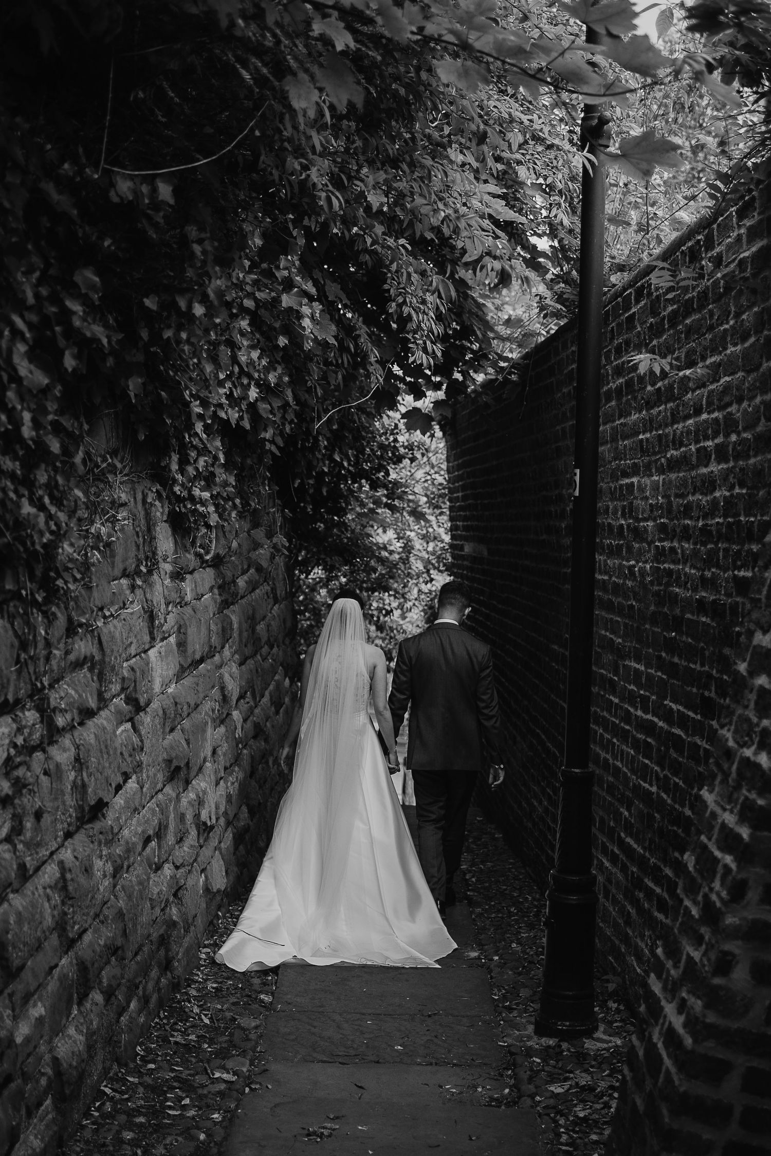 A couple walks through a narrow brick passageway covered in ivy and foliage.