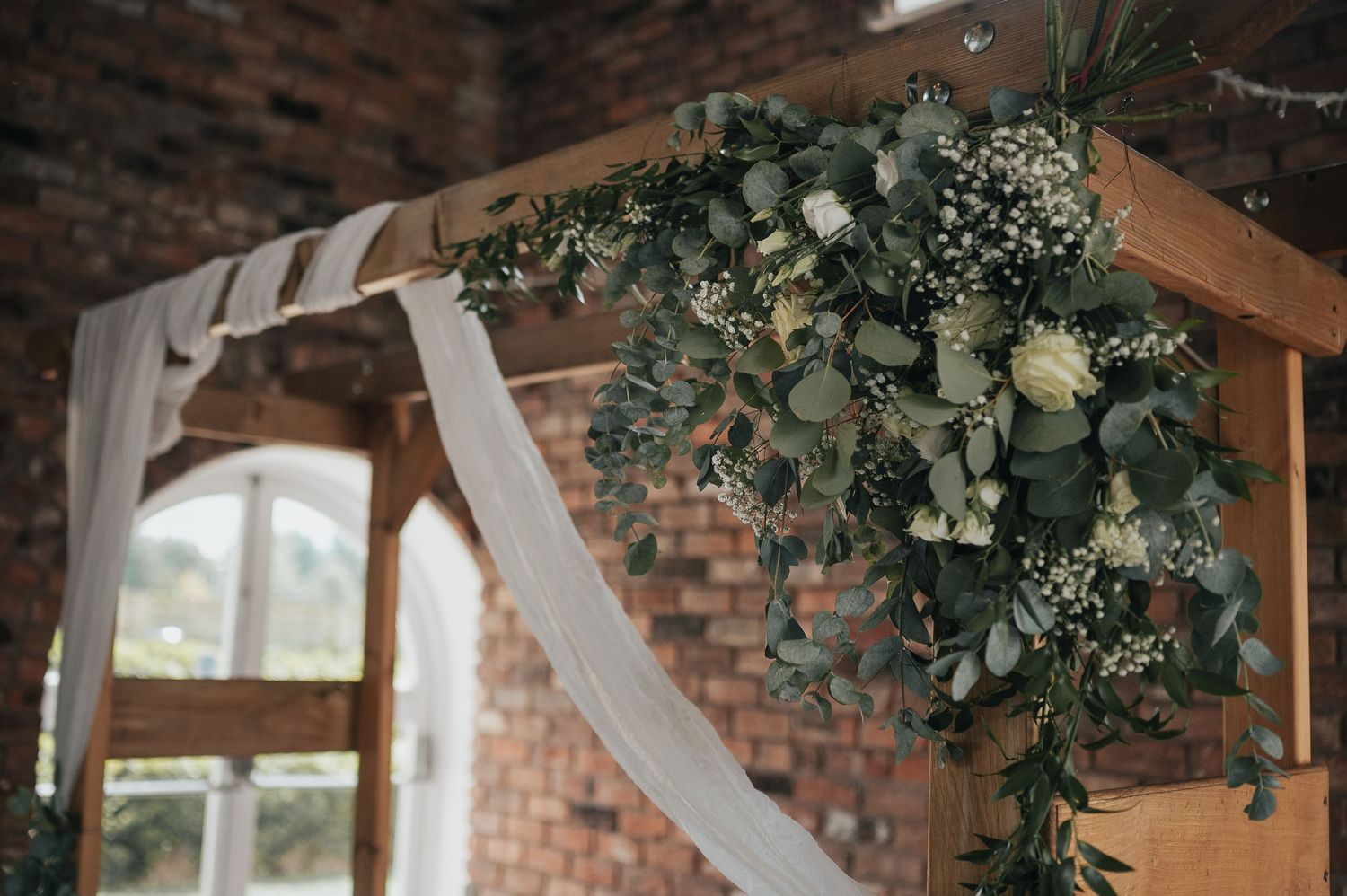Wooden wedding arch decorated with white fabric and floral arrangements against a brick wall.
