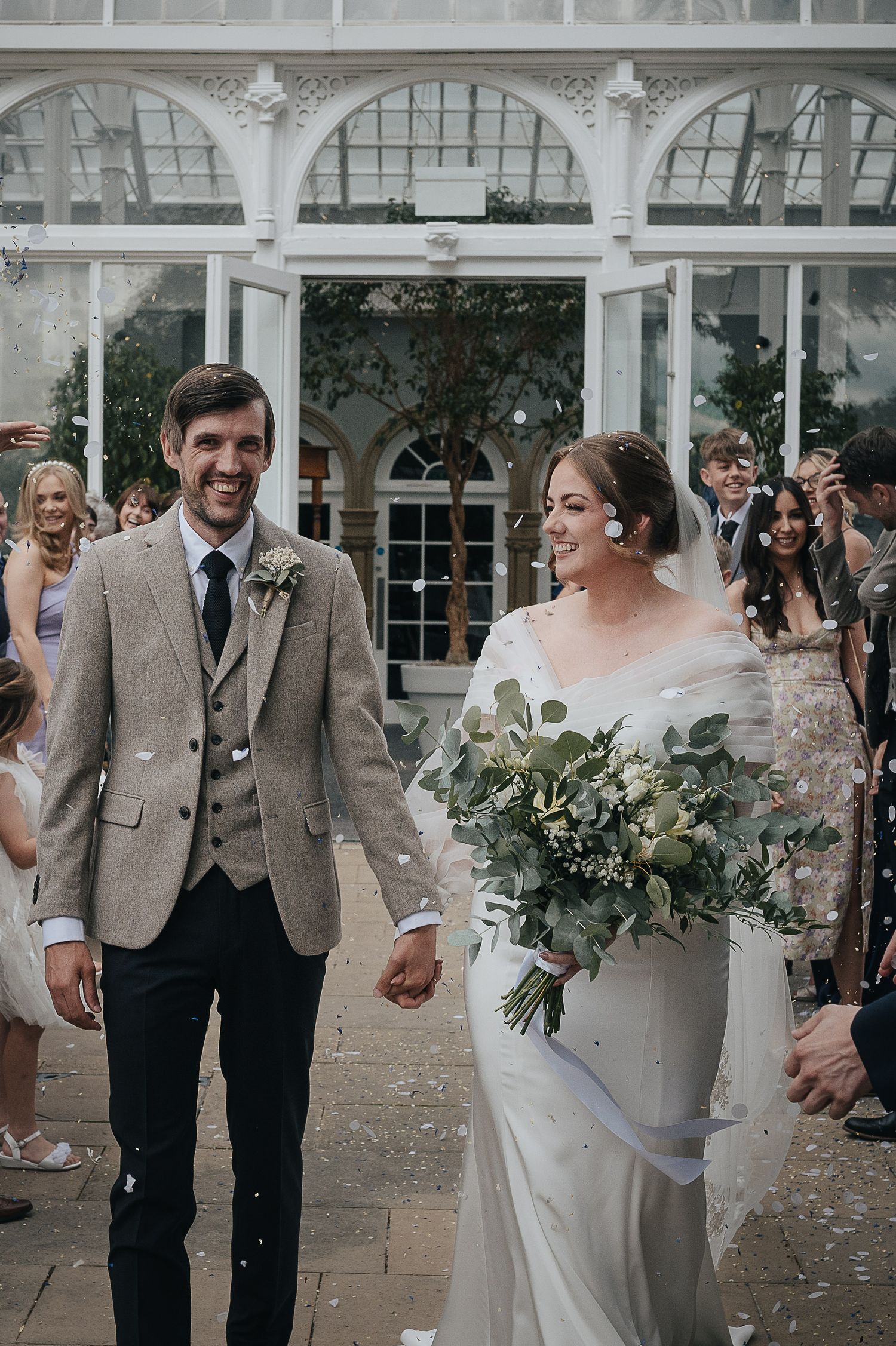 Newlyweds exit their ceremony under an ornate white conservatory while guests shower them with confetti.