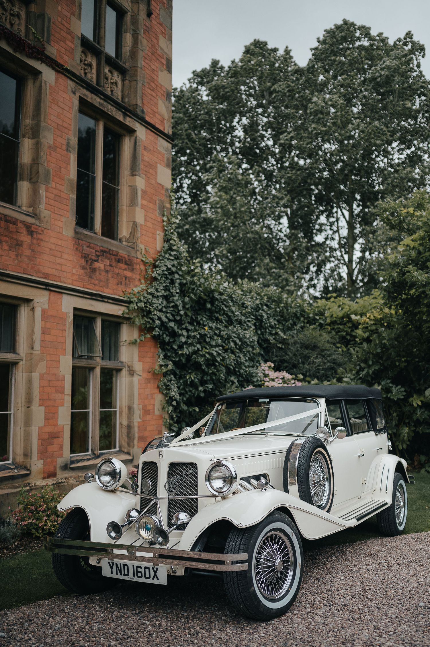 Classic white vintage Beauford wedding car parked outside red brick mansion.