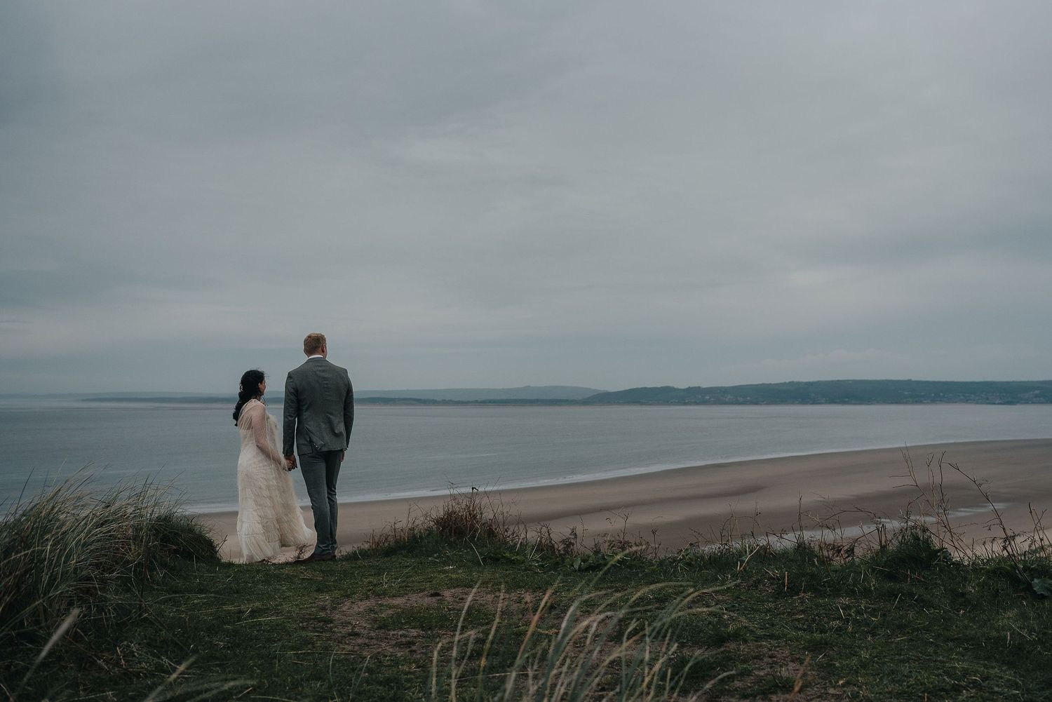 A romantic couple stands on a grassy cliff edge overlooking a vast beach landscape.