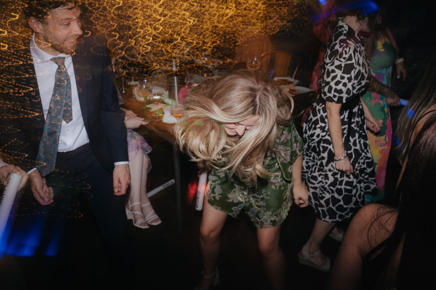 Wedding guests dancing energetically at a reception with colorful lighting.