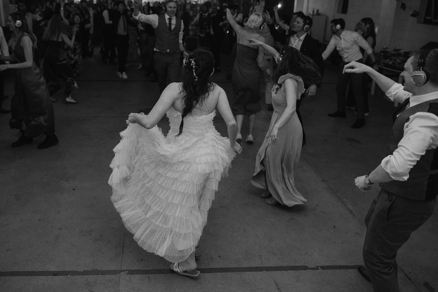 A bride in a ruffled gown dances energetically with wedding guests on the dance floor.