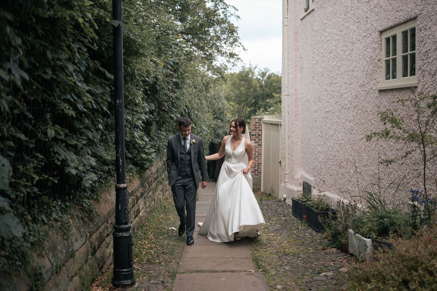 Wedding couple walking along ivy-covered garden path beside stone building.