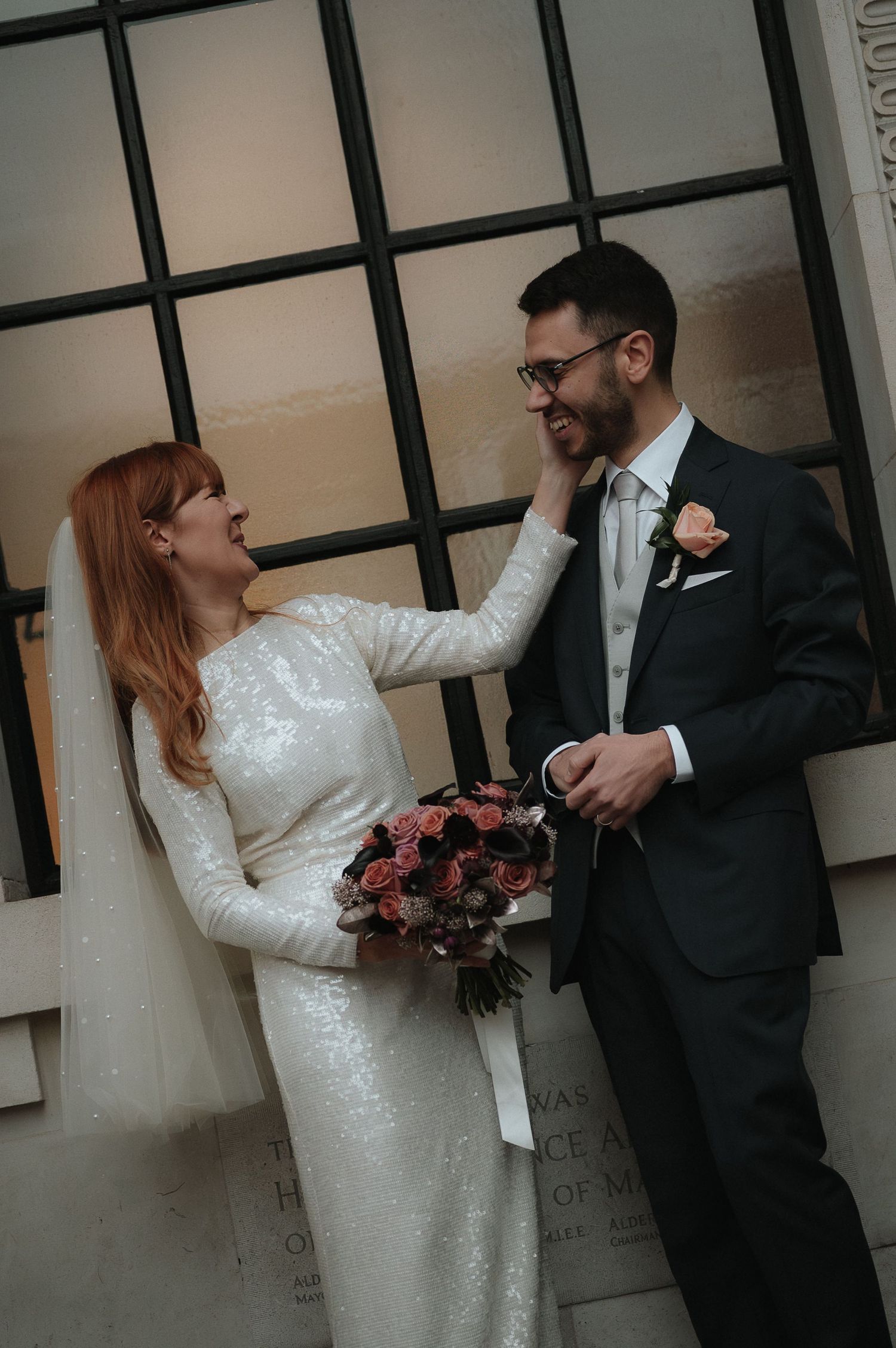 London Wedding Photographer. Bride in sequin gown touches groom's face as they share a joyful moment, holding a pink floral bouquet.