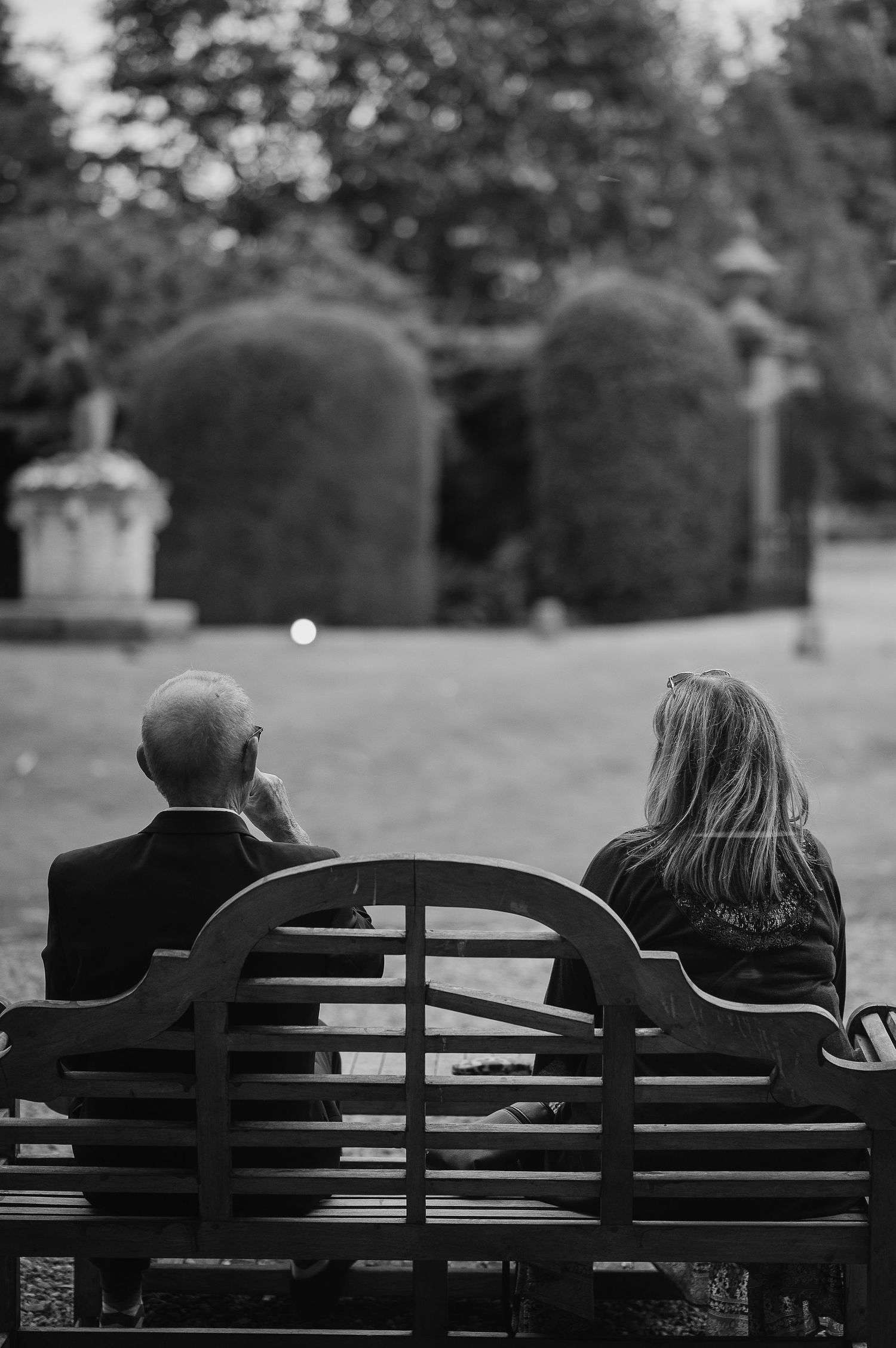 Two people share a quiet moment on a garden bench surrounded by manicured hedges.
