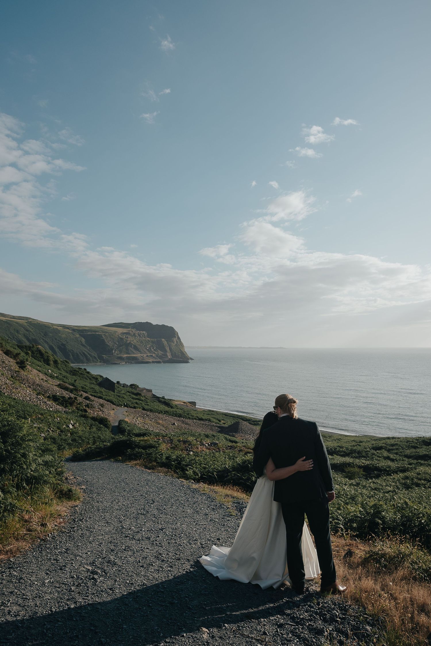Couple embracing on coastal path with dramatic cliffs and ocean view in background.