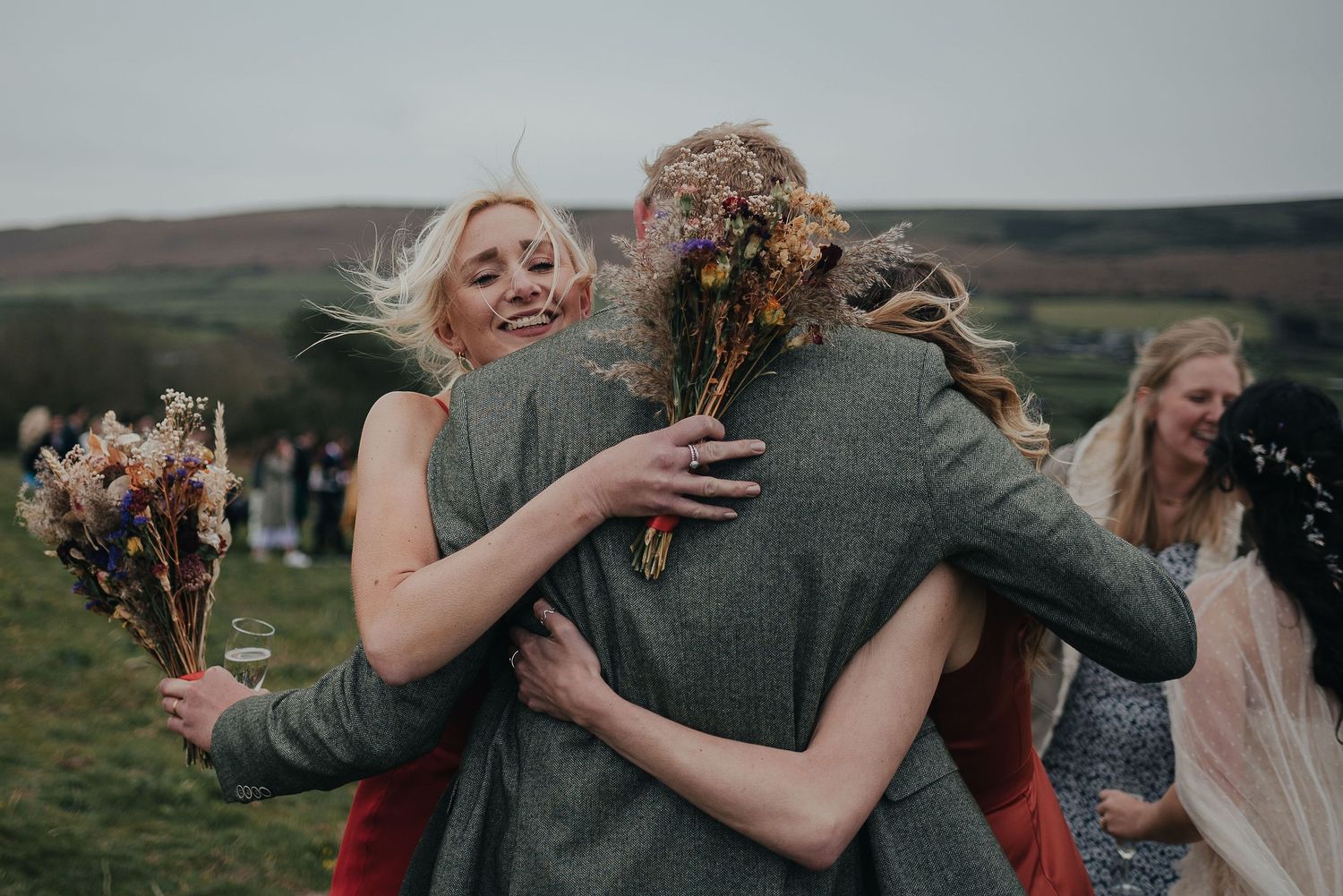 Wedding guests embrace during an outdoor celebration on green hillside.