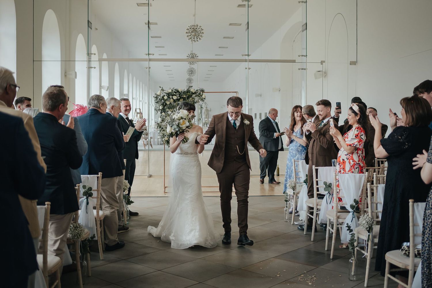 Newlyweds walk down the aisle while guests celebrate in a bright indoor venue.