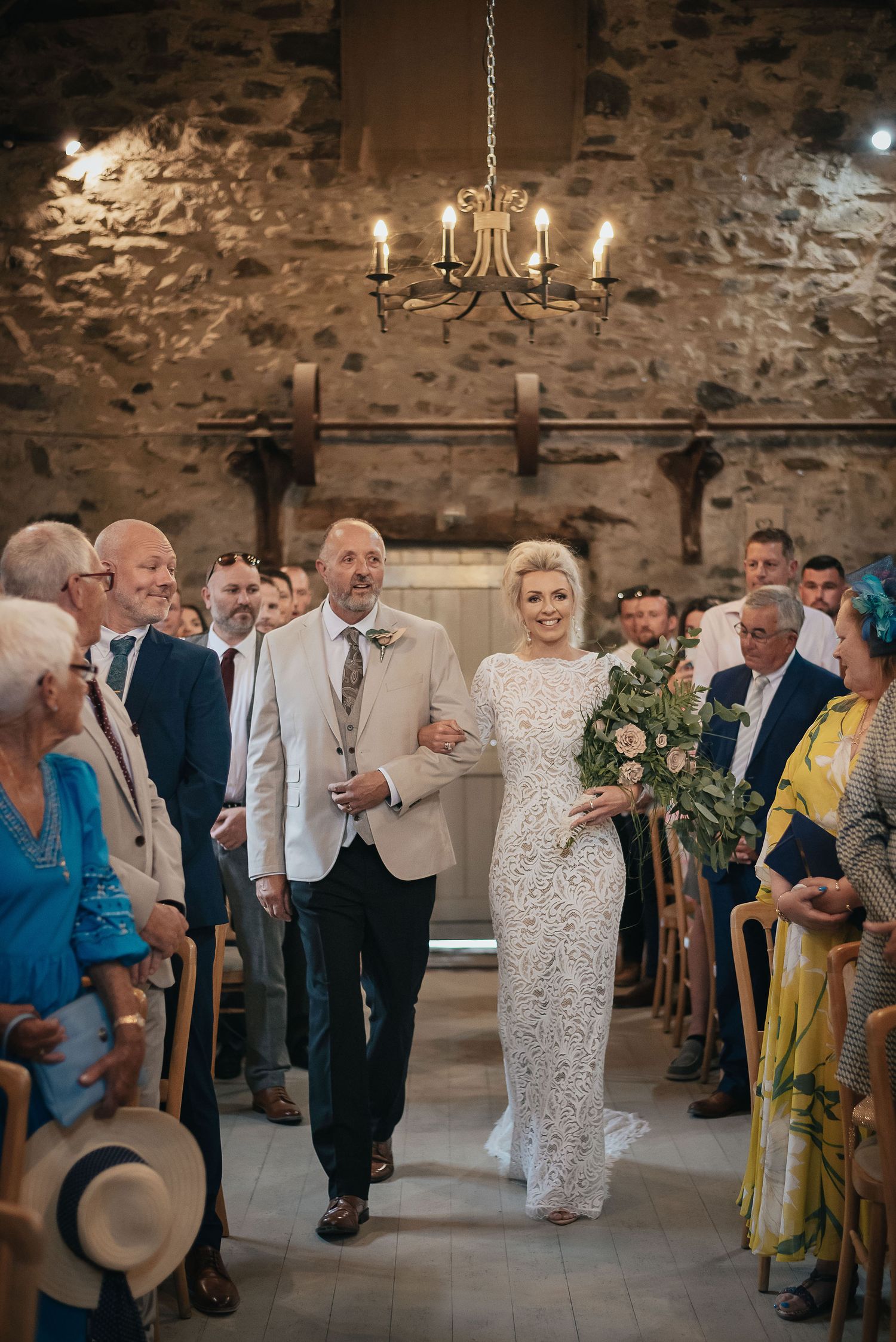 Wedding ceremony in rustic stone venue with chandelier and guests lining the aisle.
