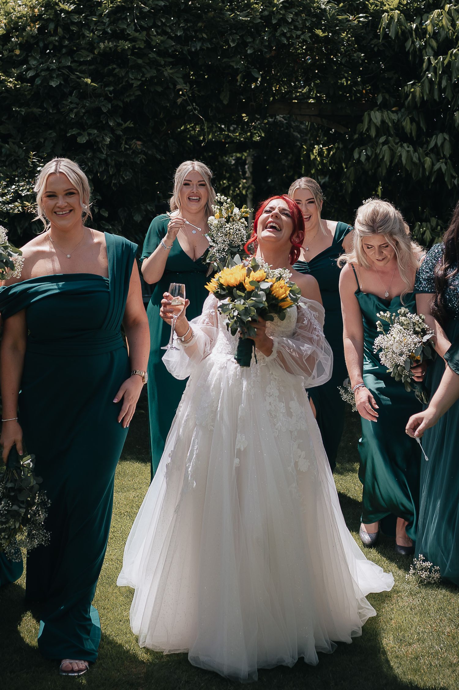 Bride in white gown laughing with bridesmaids in emerald green dresses holding sunflower bouquets.