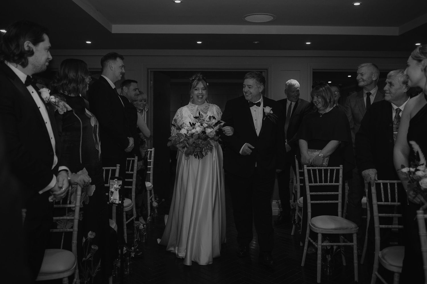 Black and white photo of wedding ceremony processional with guests standing in elegant indoor venue.