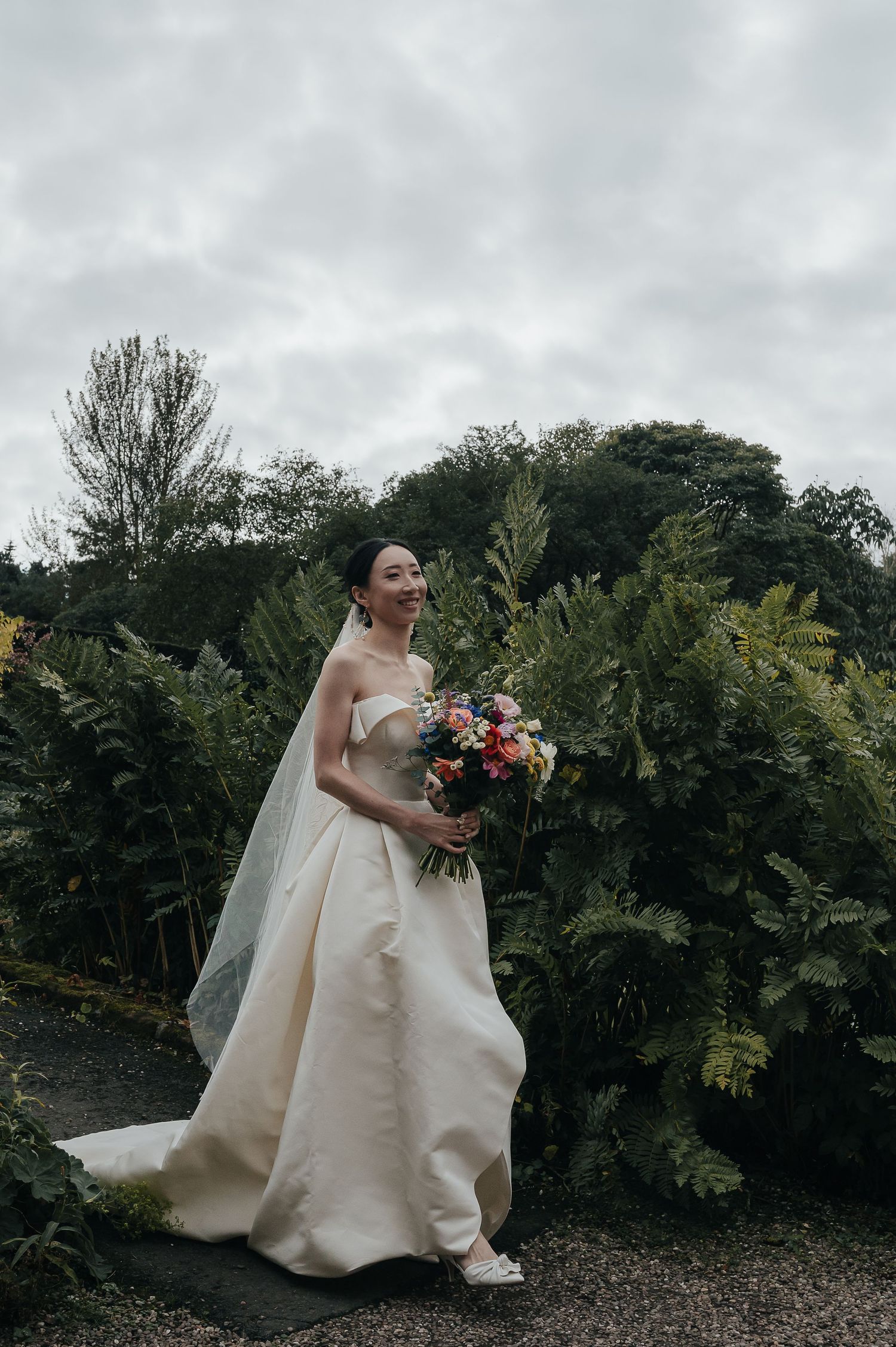 A bride in an elegant ivory wedding gown holds a vibrant bouquet against a garden backdrop on an overcast day.