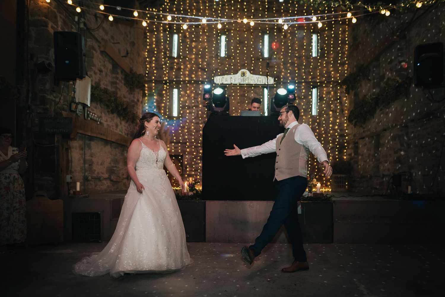 A couple dances in a rustic venue with fairy lights and brick walls creating a romantic atmosphere.