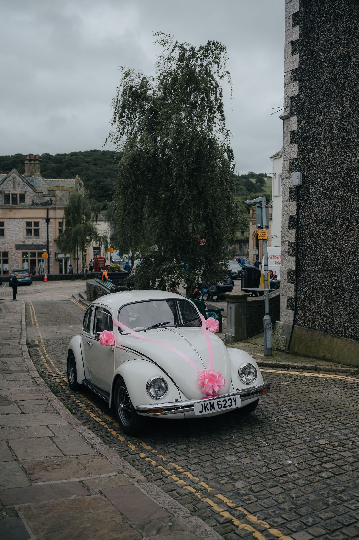 A vintage white Volkswagen Beetle decorated with pink bows parked on a cobblestone street.