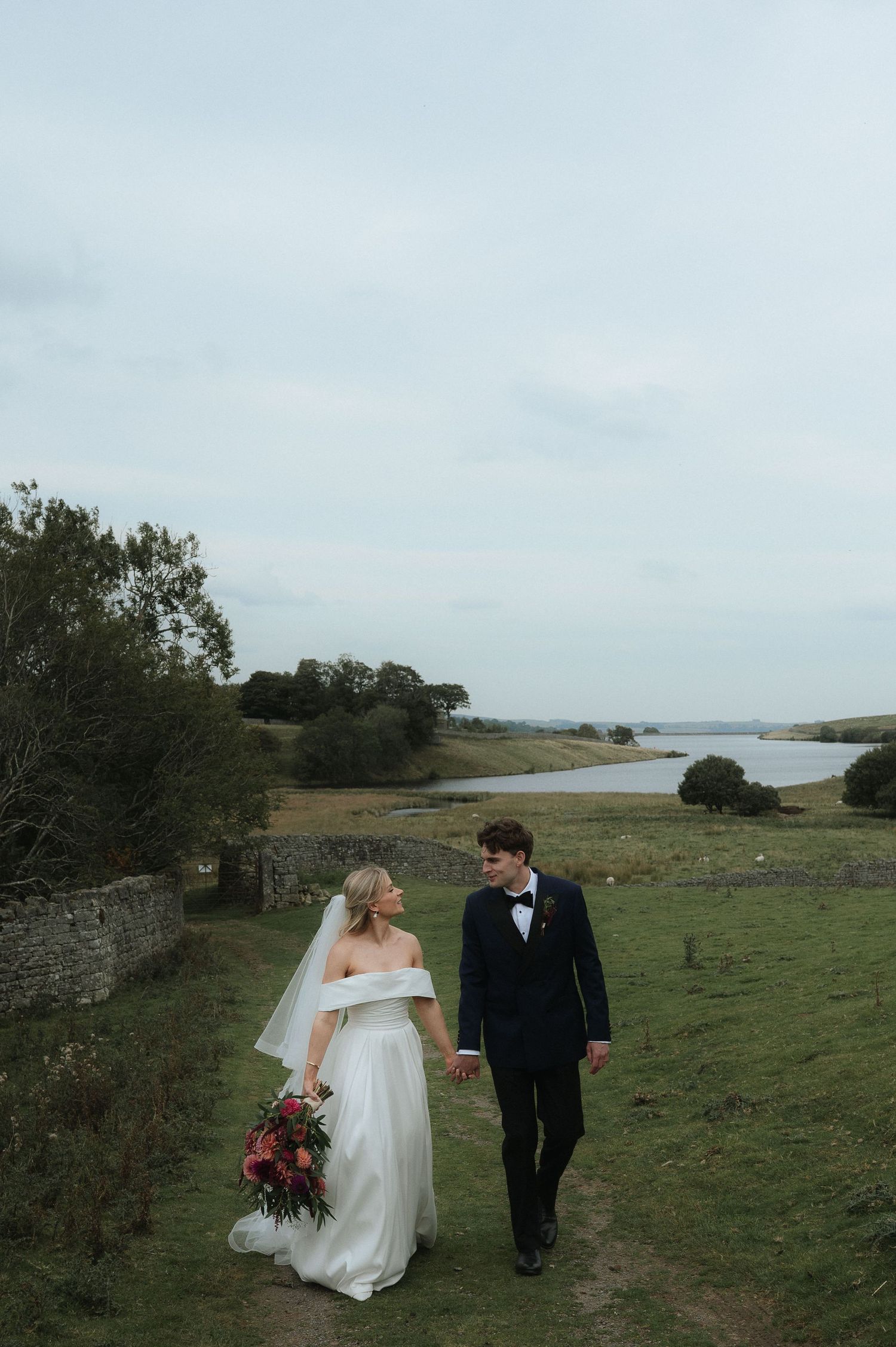 A couple walks through a scenic grassy coastal landscape on their wedding day with dramatic overcast skies.