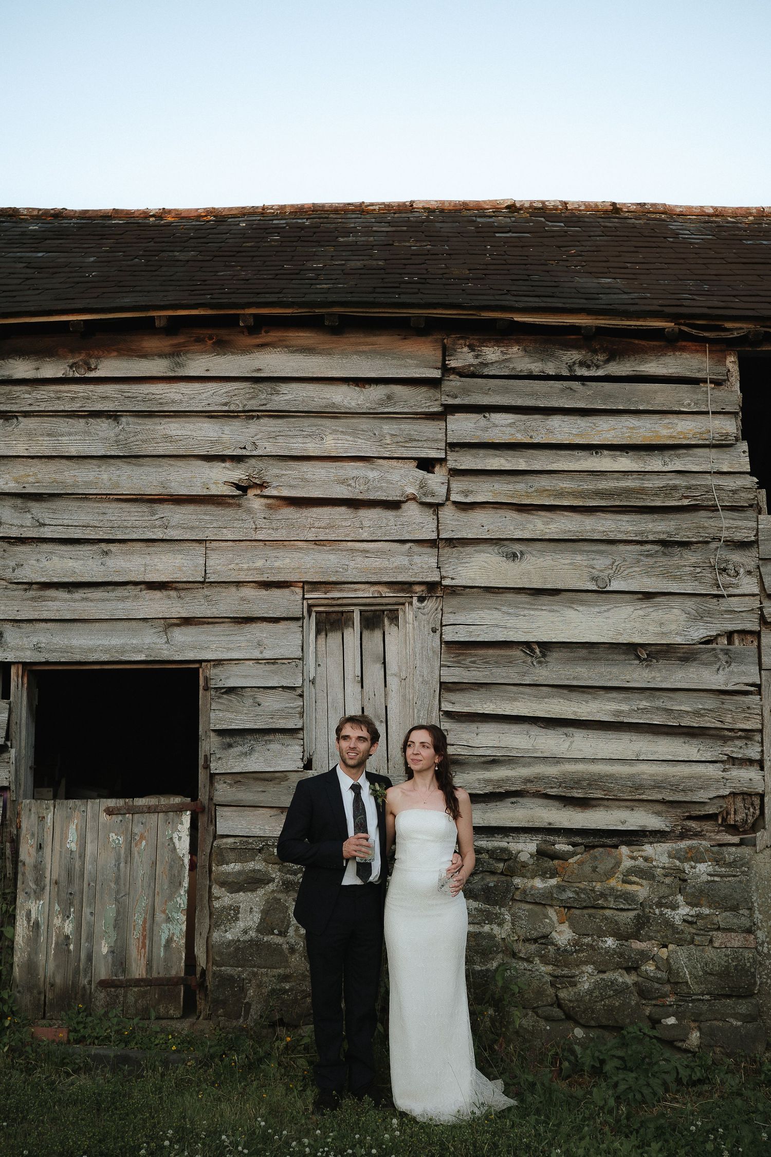 Bride and groom posing in front of a rustic weathered barn at dusk on their wedding day.
