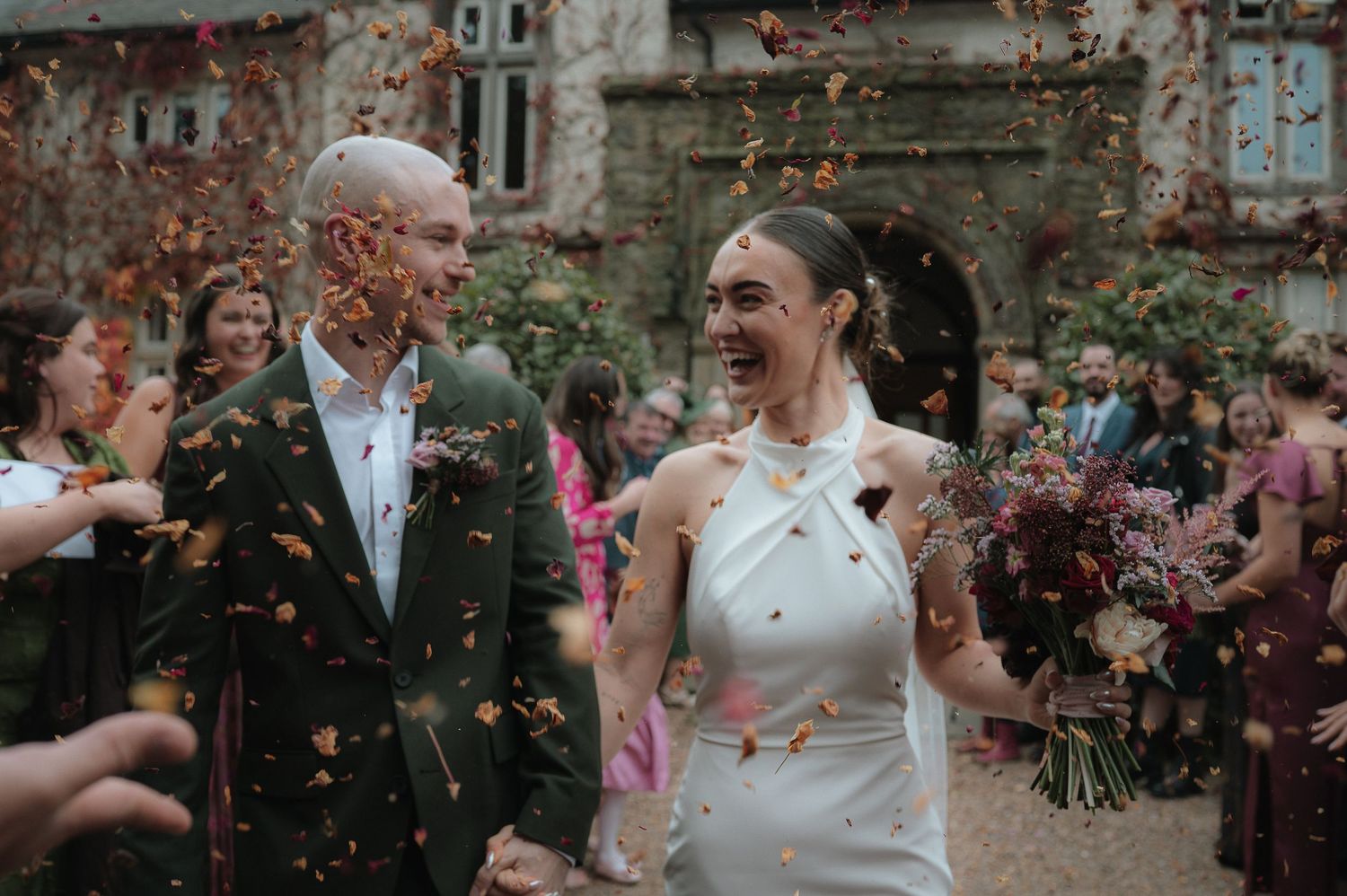 Joyful wedding celebration with guests throwing pink flower petals at a couple outside a stone building.