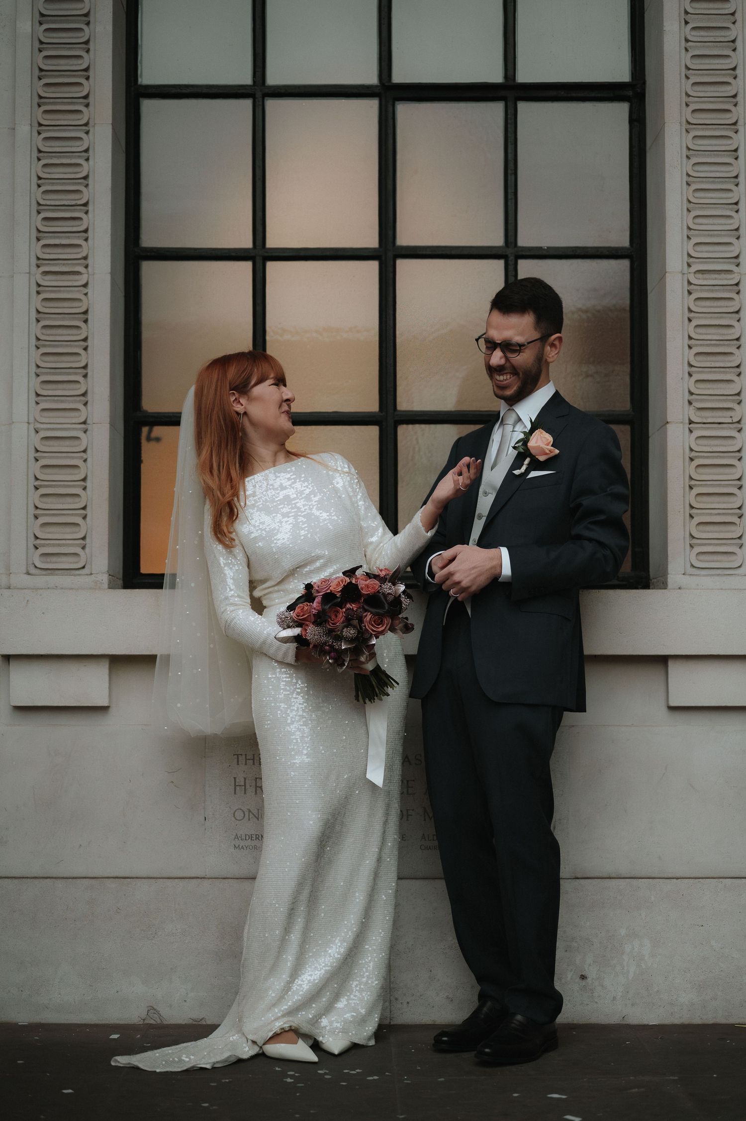 Bride in white sequin gown holding bouquet stands with groom in dark suit before large window. London Wedding Photographer.