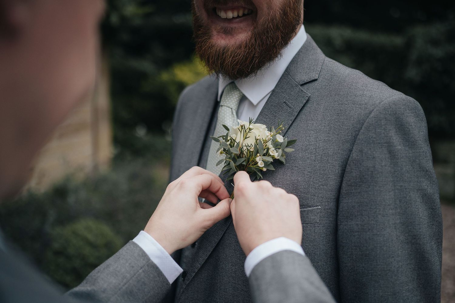 Close-up of hands pinning a white boutonniere on a grey suit lapel.
