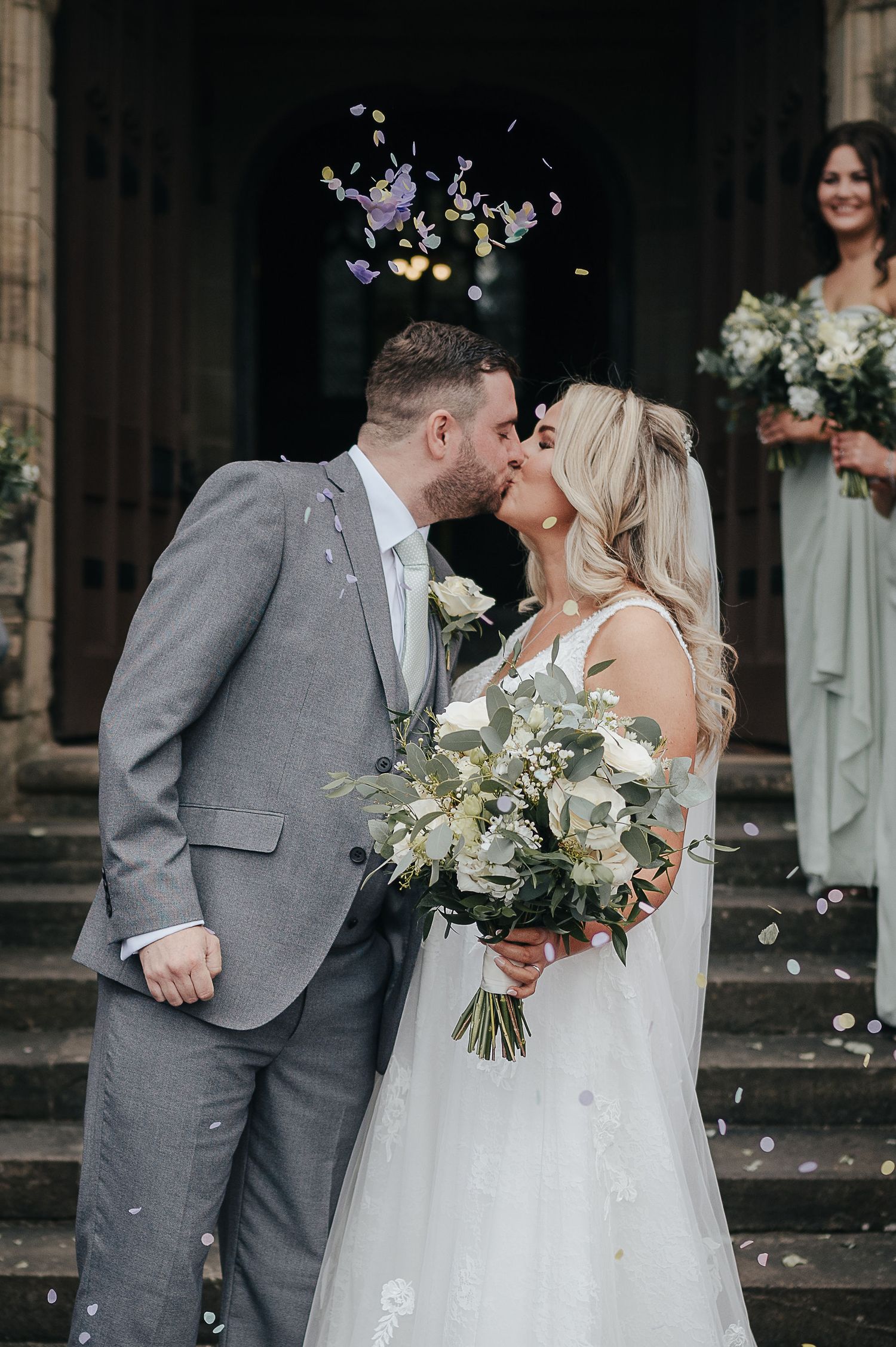 Newlyweds share a kiss on stone steps as confetti falls around them during an elegant outdoor wedding ceremony.