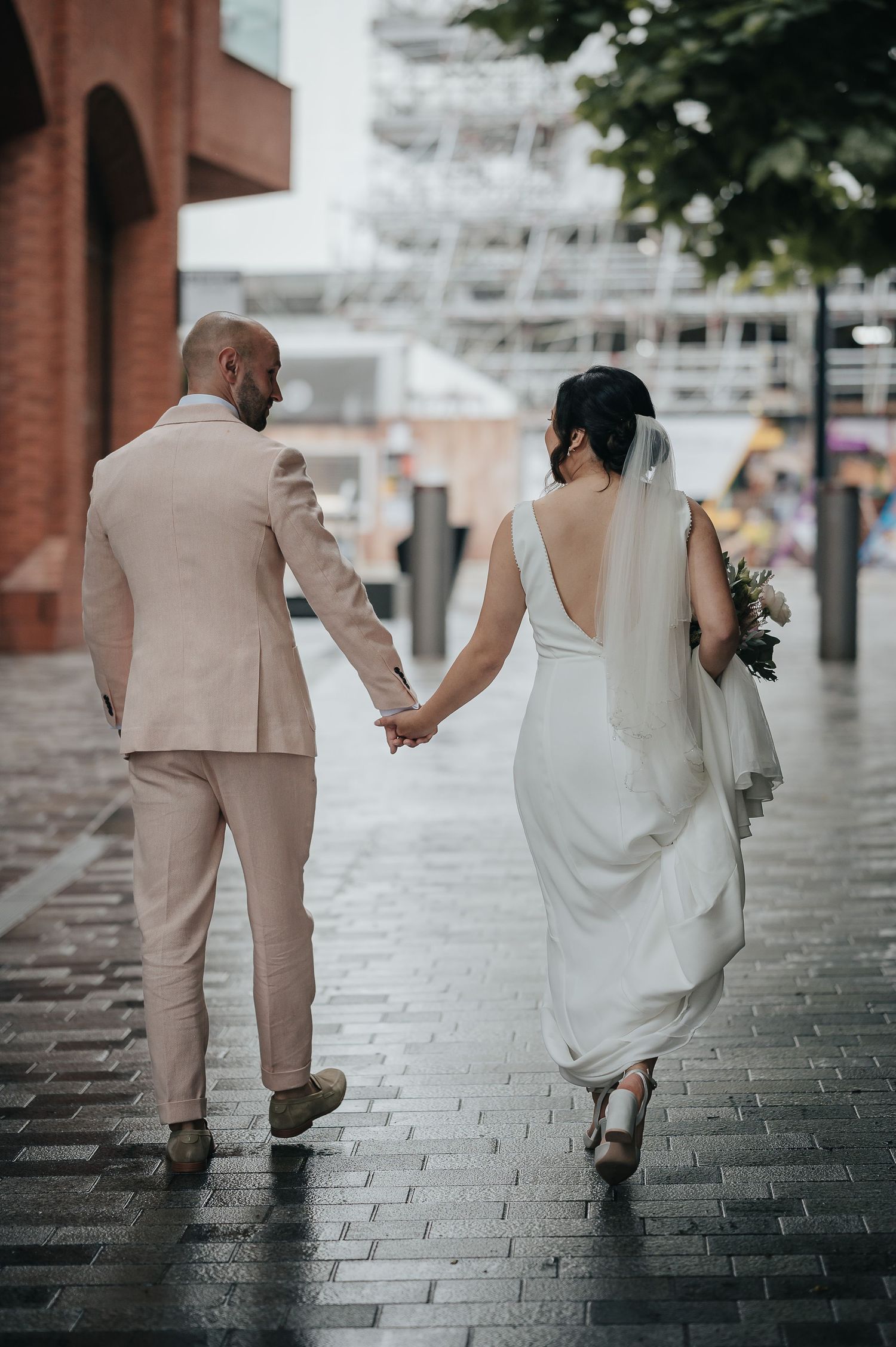 Newlyweds holding hands while walking on a rainy cobblestone street in formal attire.