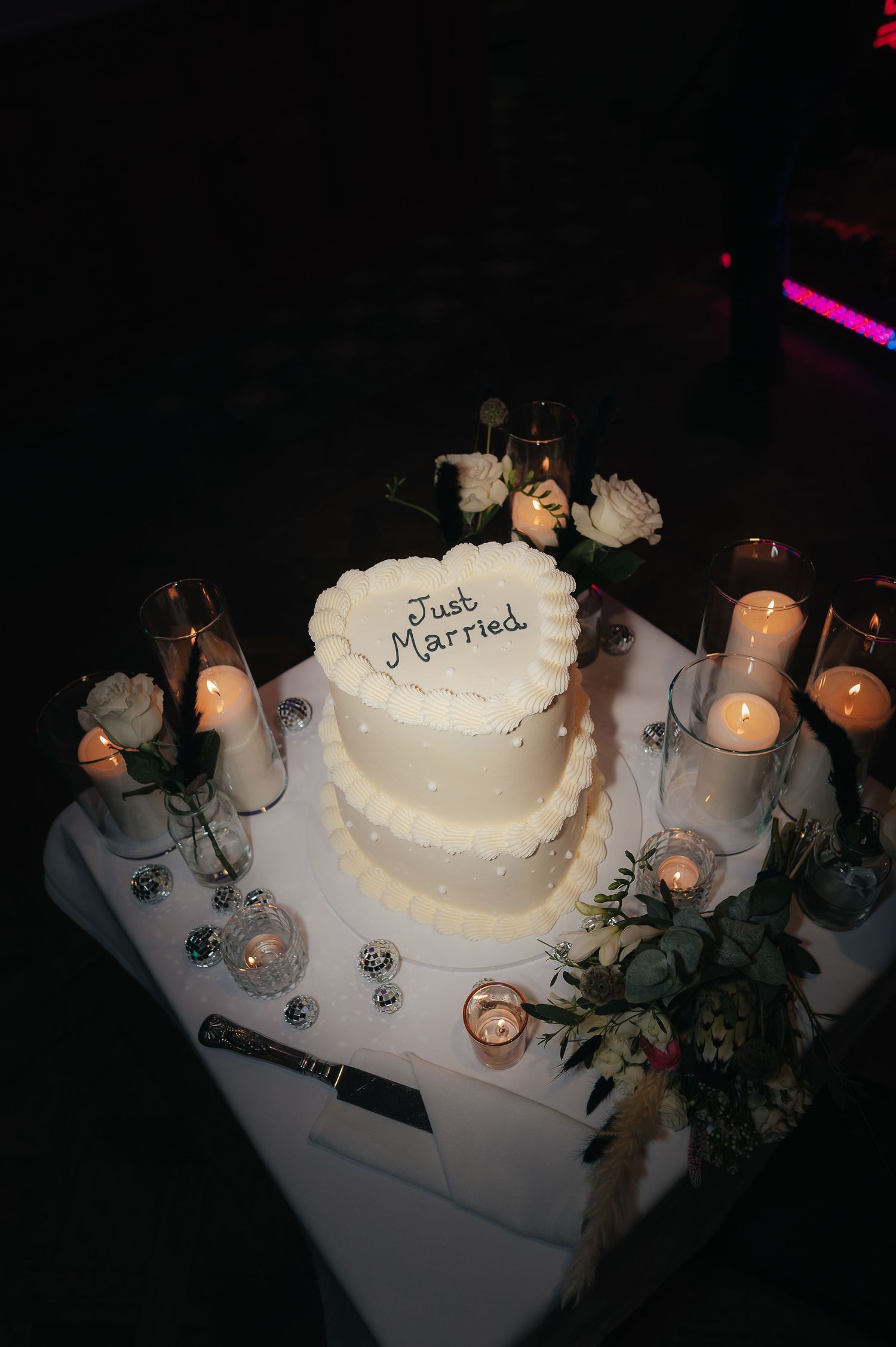 A white two-tiered wedding cake surrounded by lit candles and floral decorations on an elegant table setting.