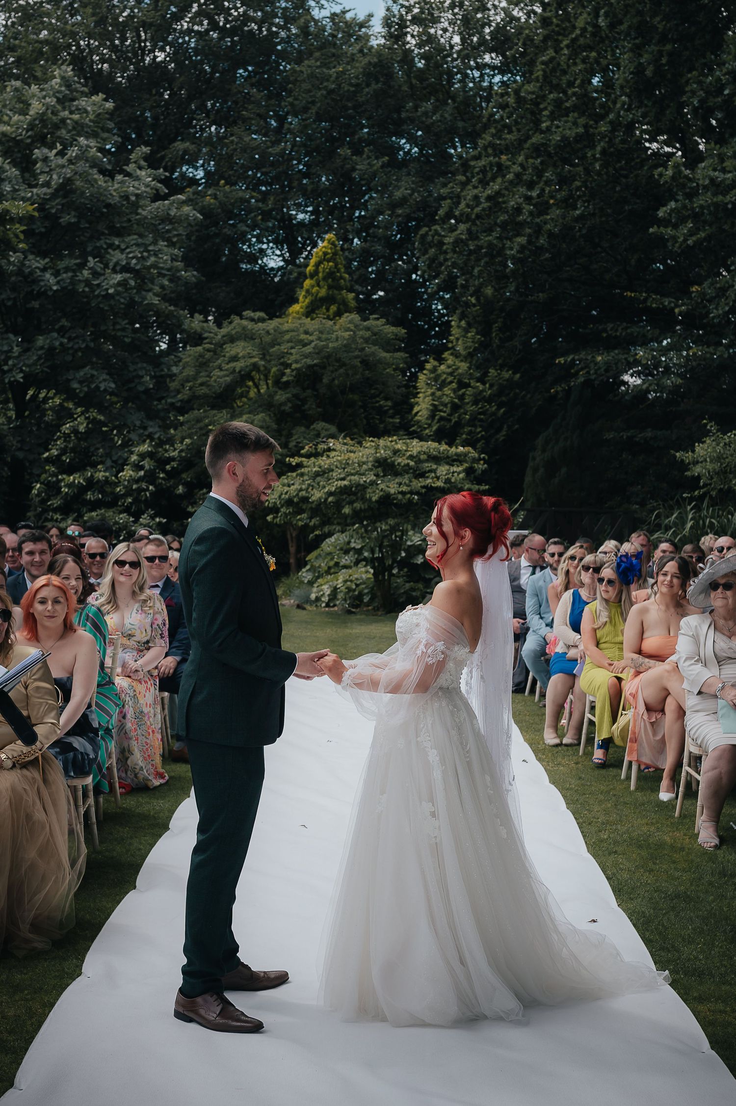 Wedding ceremony in progress on white aisle runner surrounded by seated guests in garden setting.