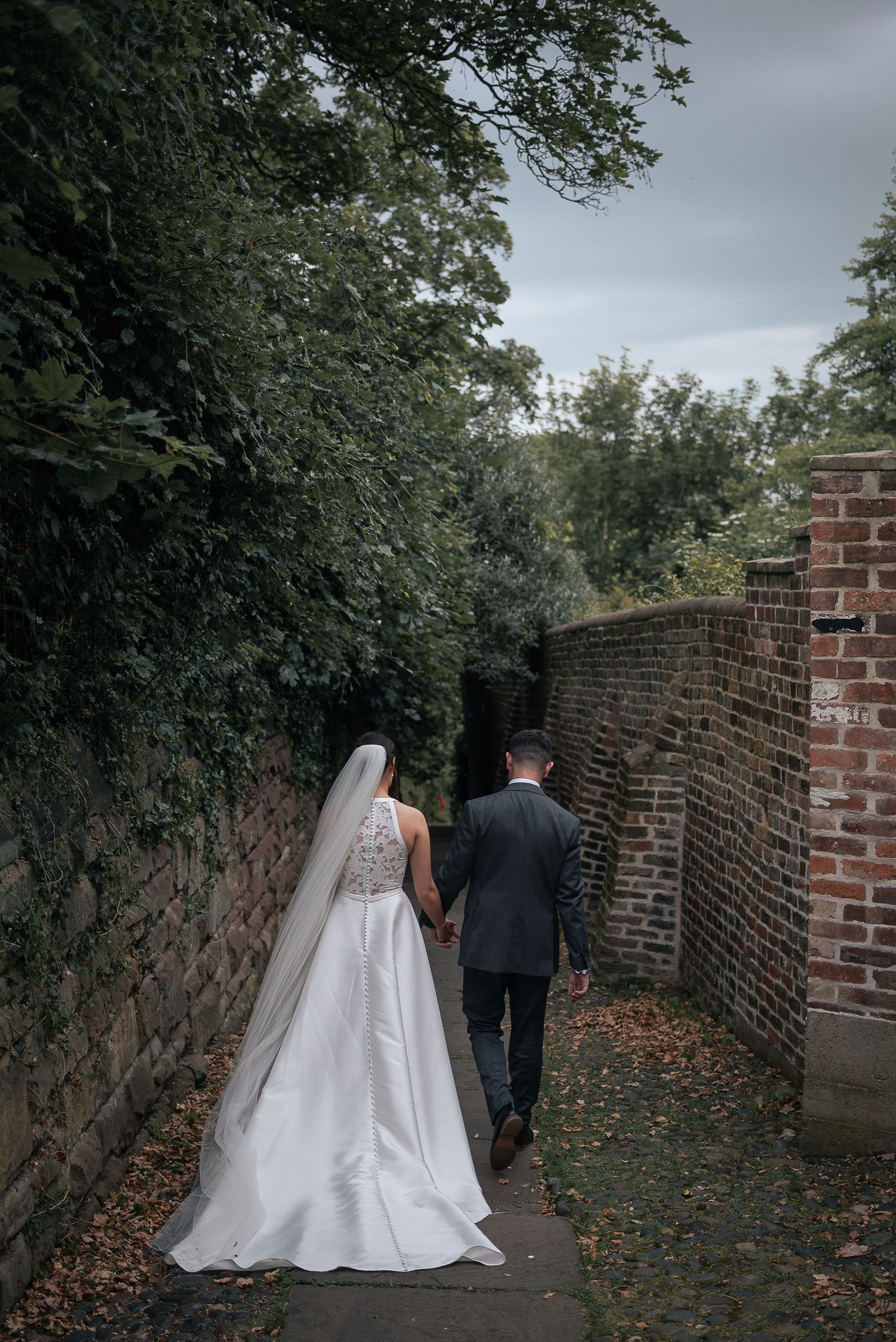 Couple walking along brick-walled garden path in wedding attire.