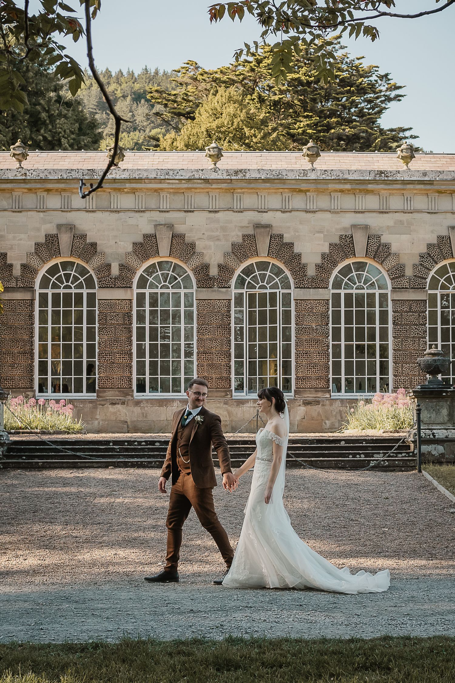 Couple walking near historic stone building with arched windows during sunset wedding photoshoot.