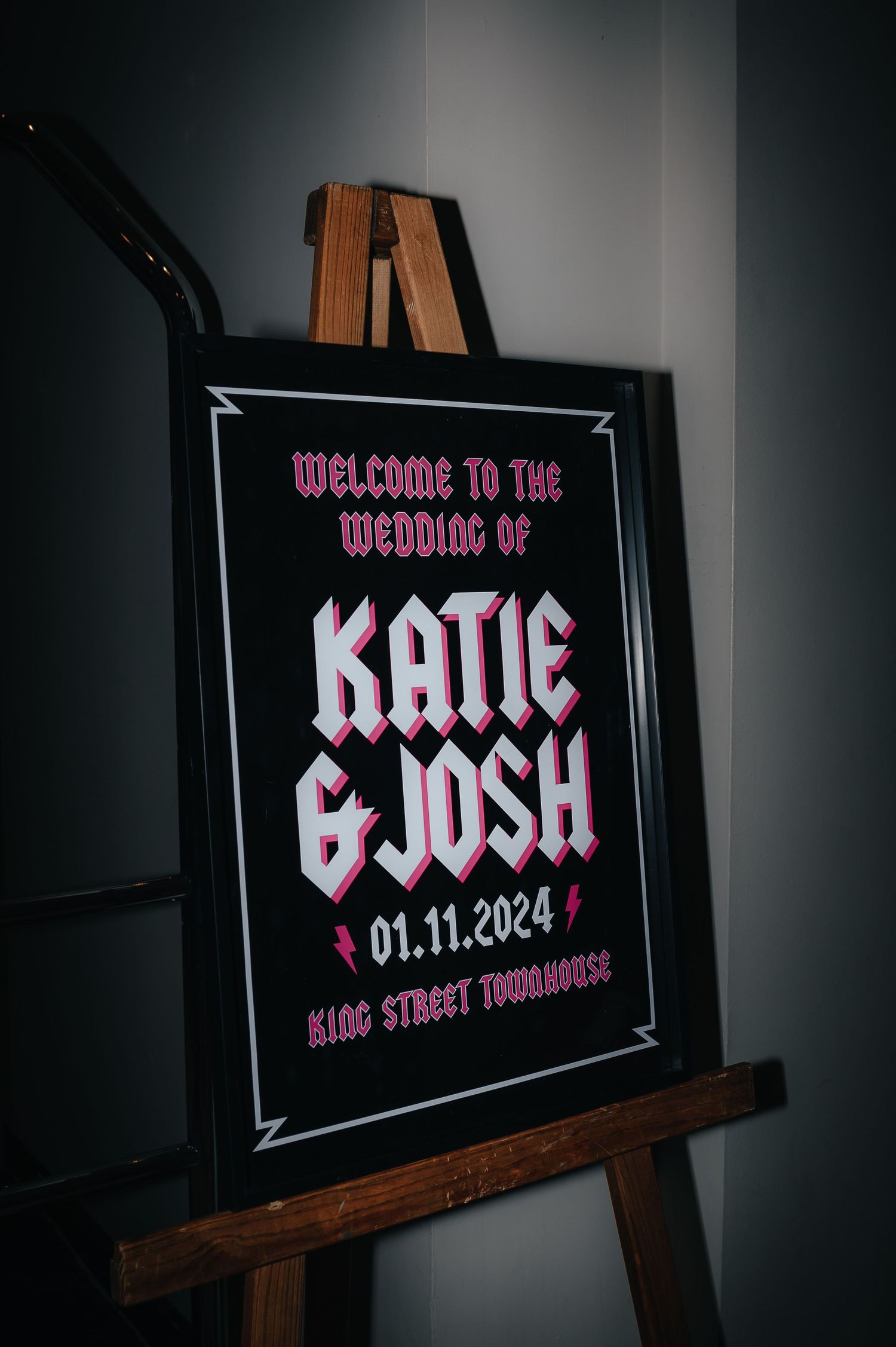 Wedding welcome sign on wooden easel displaying 'Katie & Josh' with pink text and date 01.11.2024 against black background.