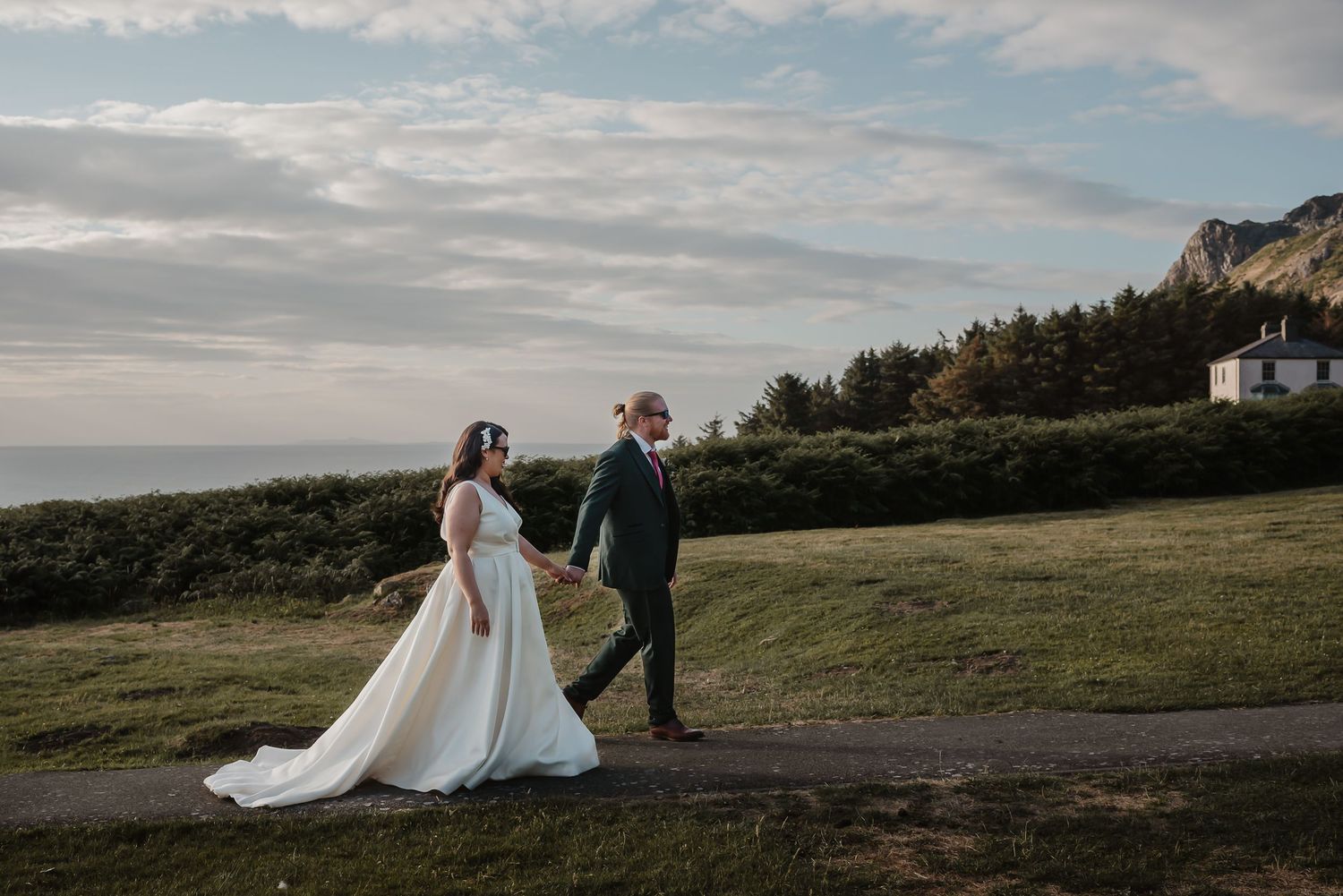 Newlyweds walking along coastal path with dramatic cloudy sky and cliffs in background.