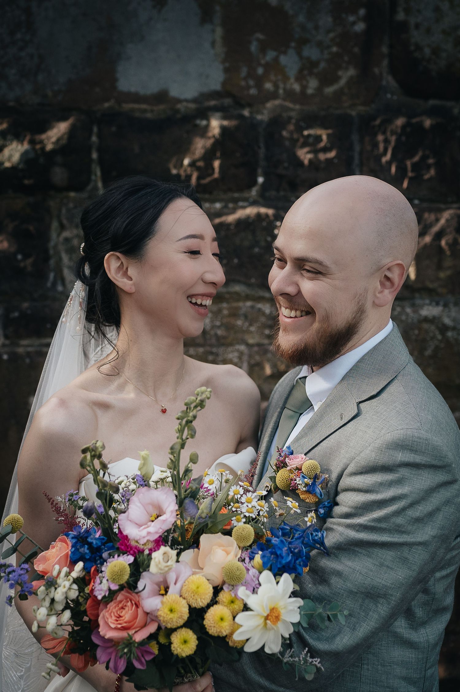Happy newlyweds share a candid moment while holding vibrant multicolored wedding bouquet.