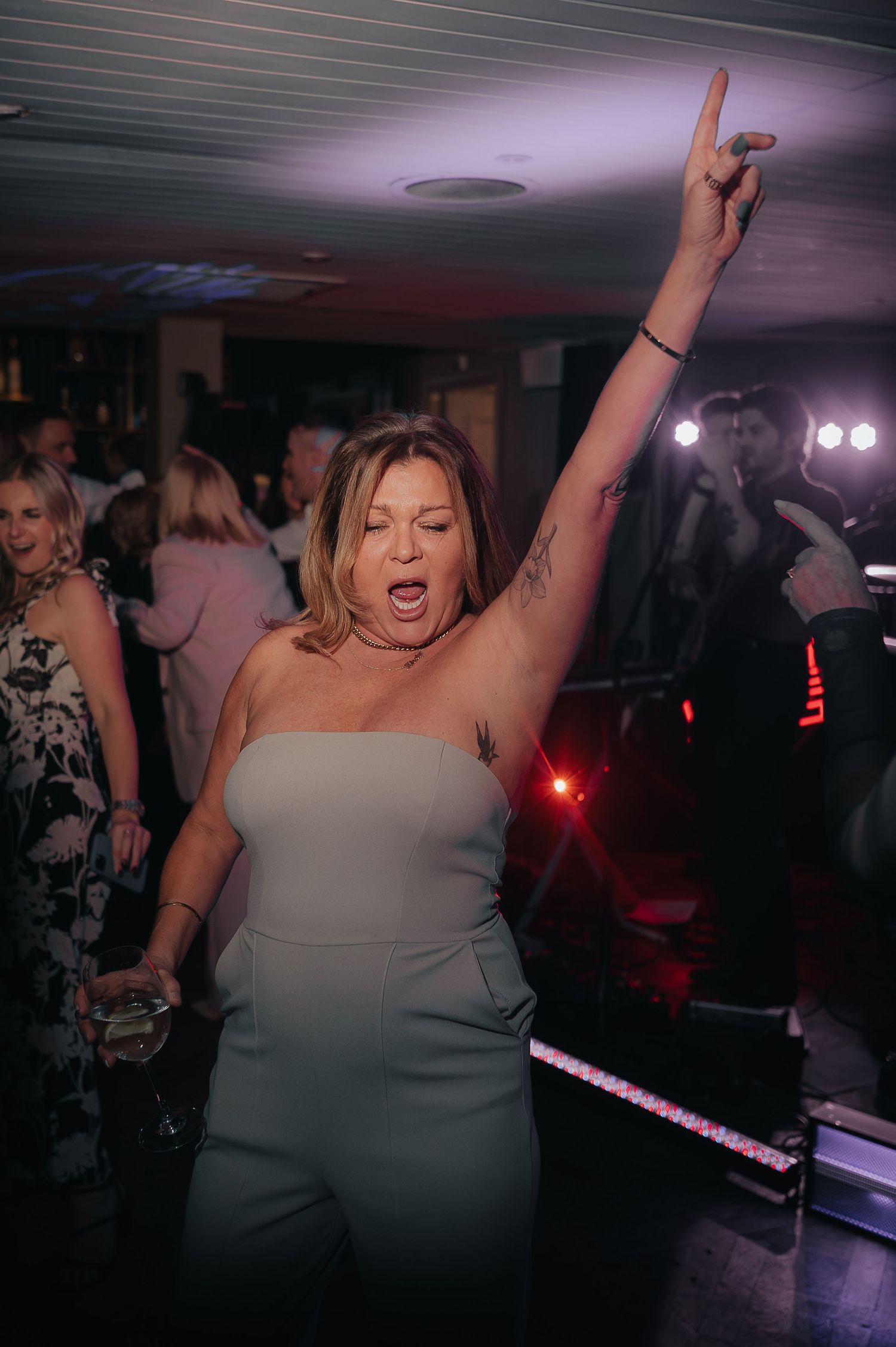 A figure in a white dress energetically dances at a party under dim lighting with upraised arms.