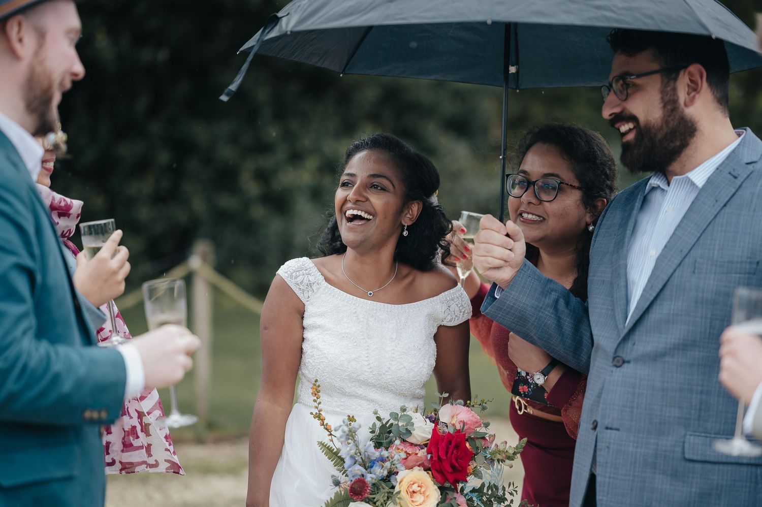 Wedding party sharing laughs under an umbrella during outdoor reception.
