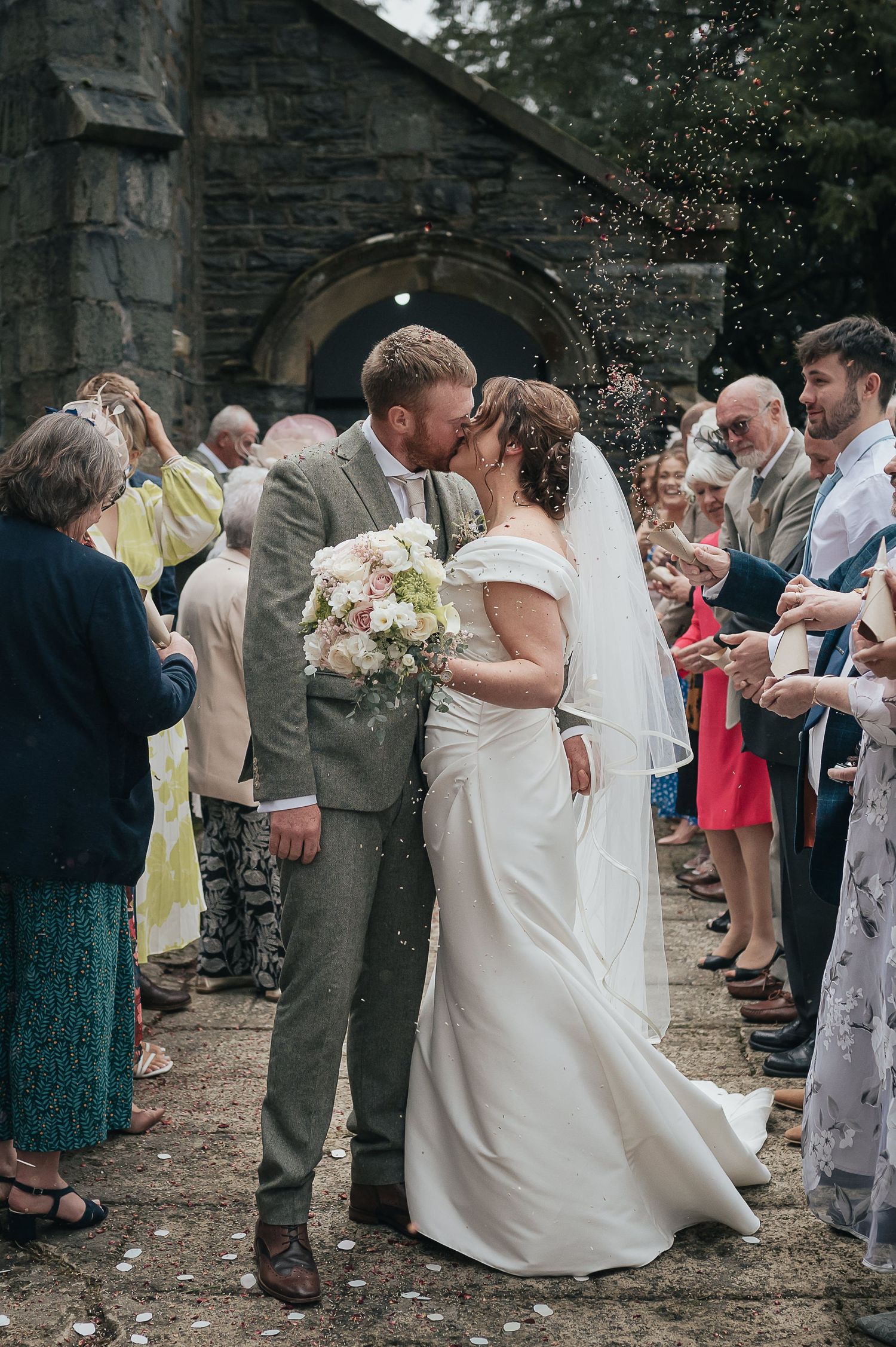 Wedding celebration outside stone archway with guests throwing confetti.
